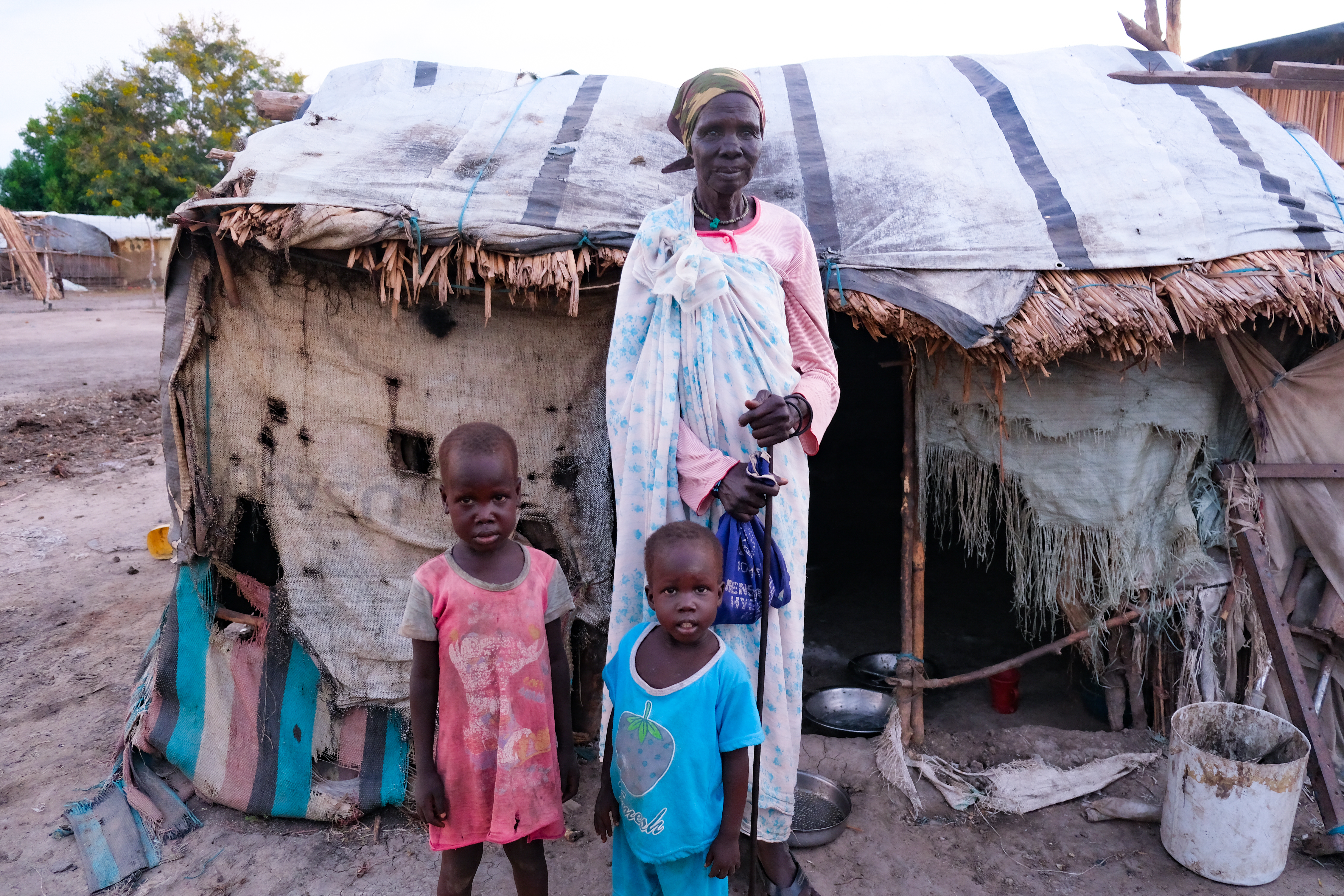 Mary Nyashin Tsief (70) stands in front of her home in Rubkona camp for people displaced by flooding and violent conflict. She is accompanied by her two grandchildren, Nyamen Iuok Ram (age 5) and Nyawich Iuok Ram (age 3), who live in a tent adjacent to hers.