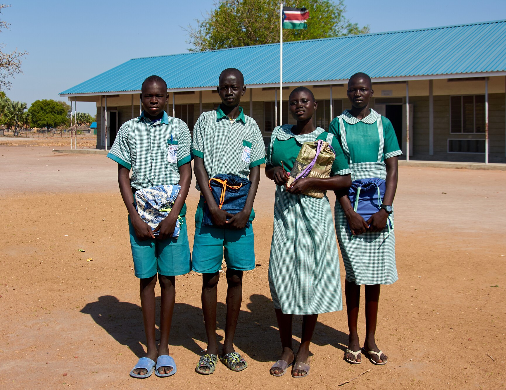 Two boys and two girls stand outside of a school holding school kits