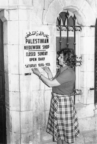 A black and white photo of a woman hanging a sign that reads "Palestinian Needle Shop Closed Sunday Open Daily Saturday 10-4" 