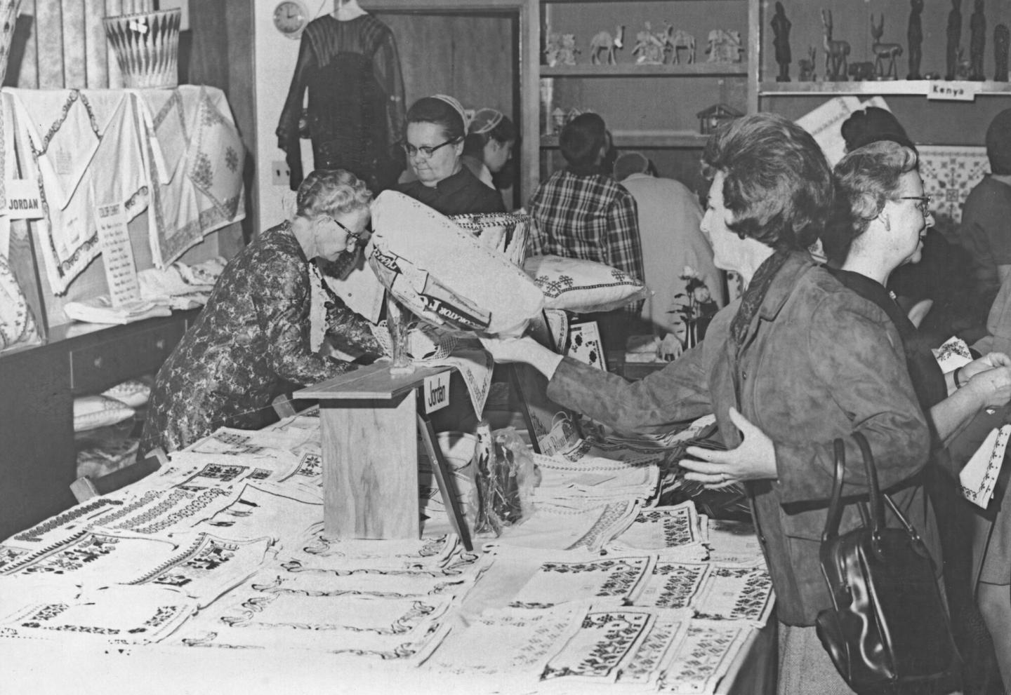 A black and white photo of women shopping at a craft store with Palestinian sewing on display