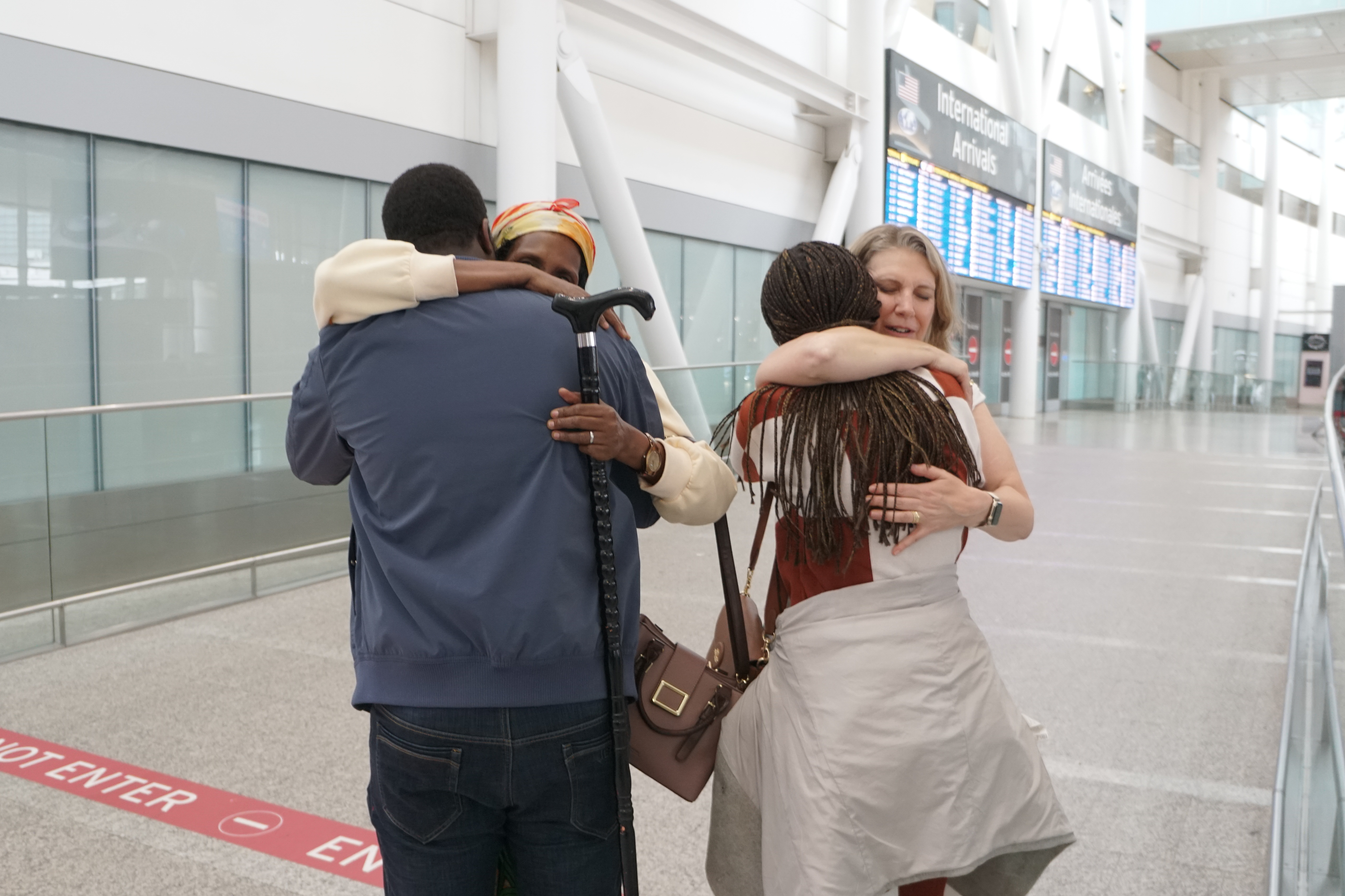 Two sets of people are embracing in an airport arrivals area.