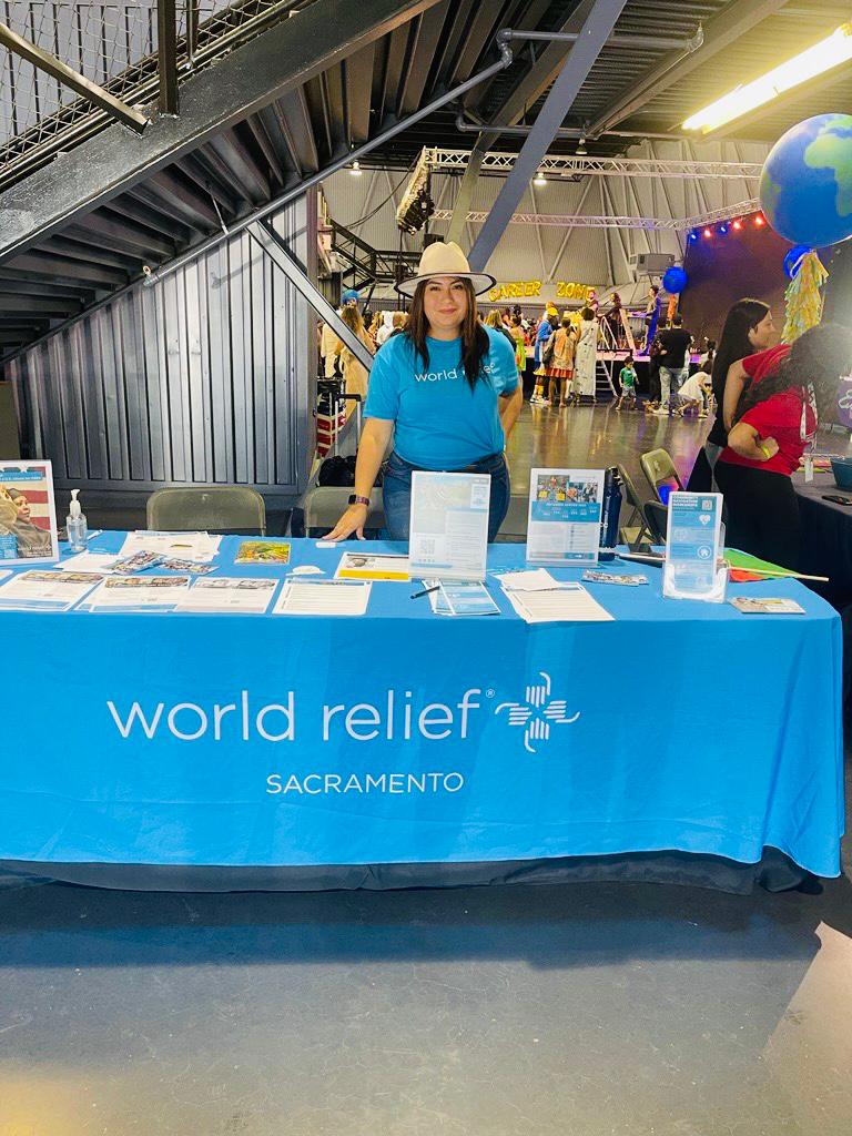 A person stands behind a table that reads "World Relief Sacramento" and is covered in covered informative documents and brochures. Their shirt is for the same organization.