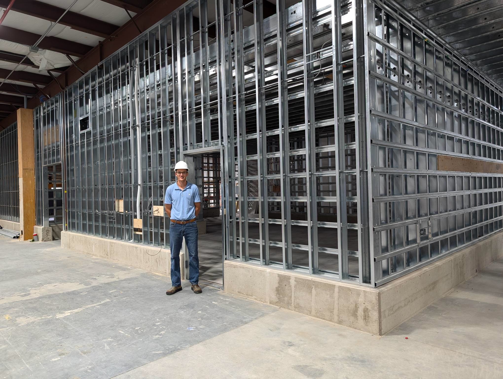 man in hard hat stands in front of a wall of steel studs under construction