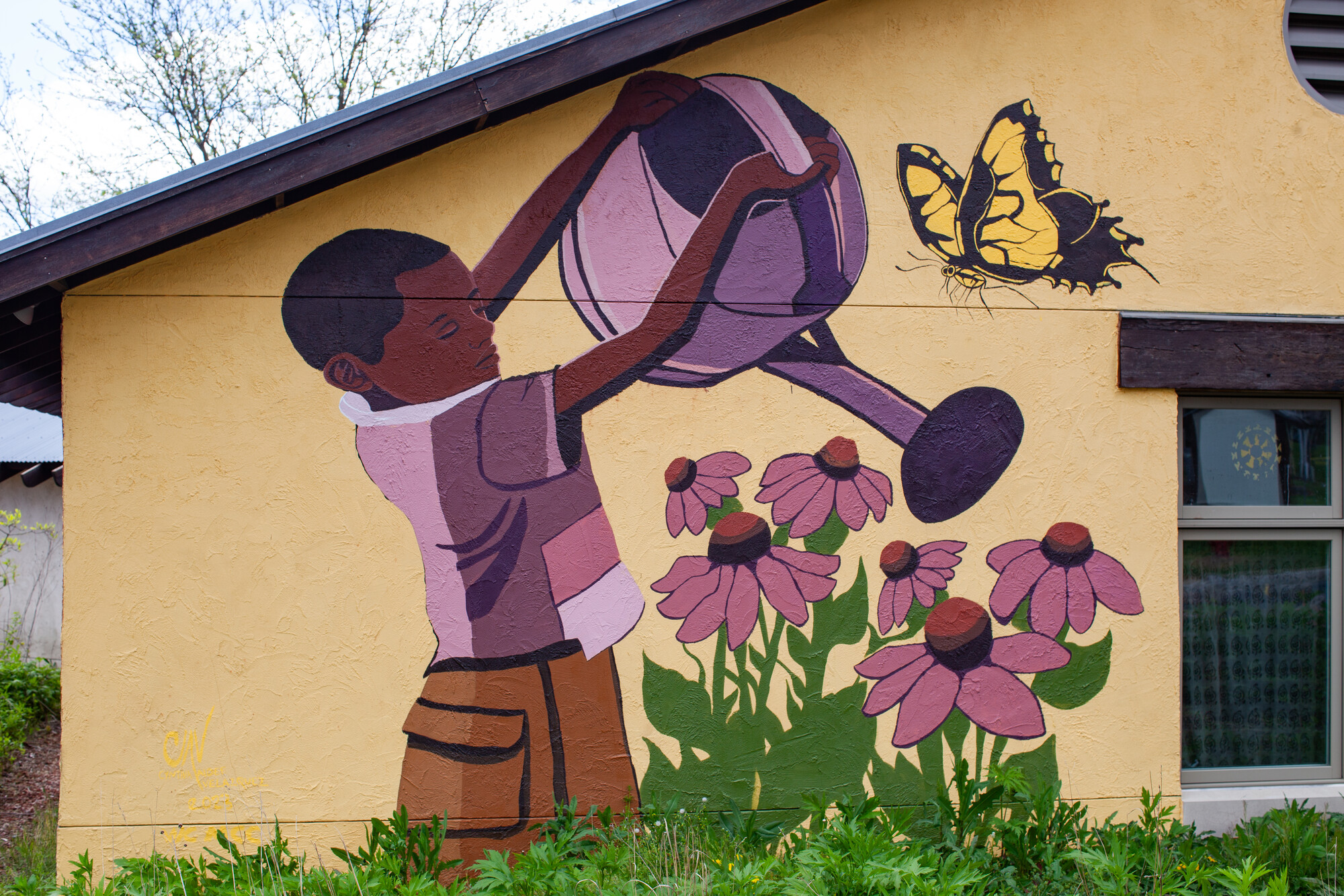 A mural depicts a child watering flowers with a large butterfly nearby. The artwork is on a building's wall, with a clear sky above and grass below.
