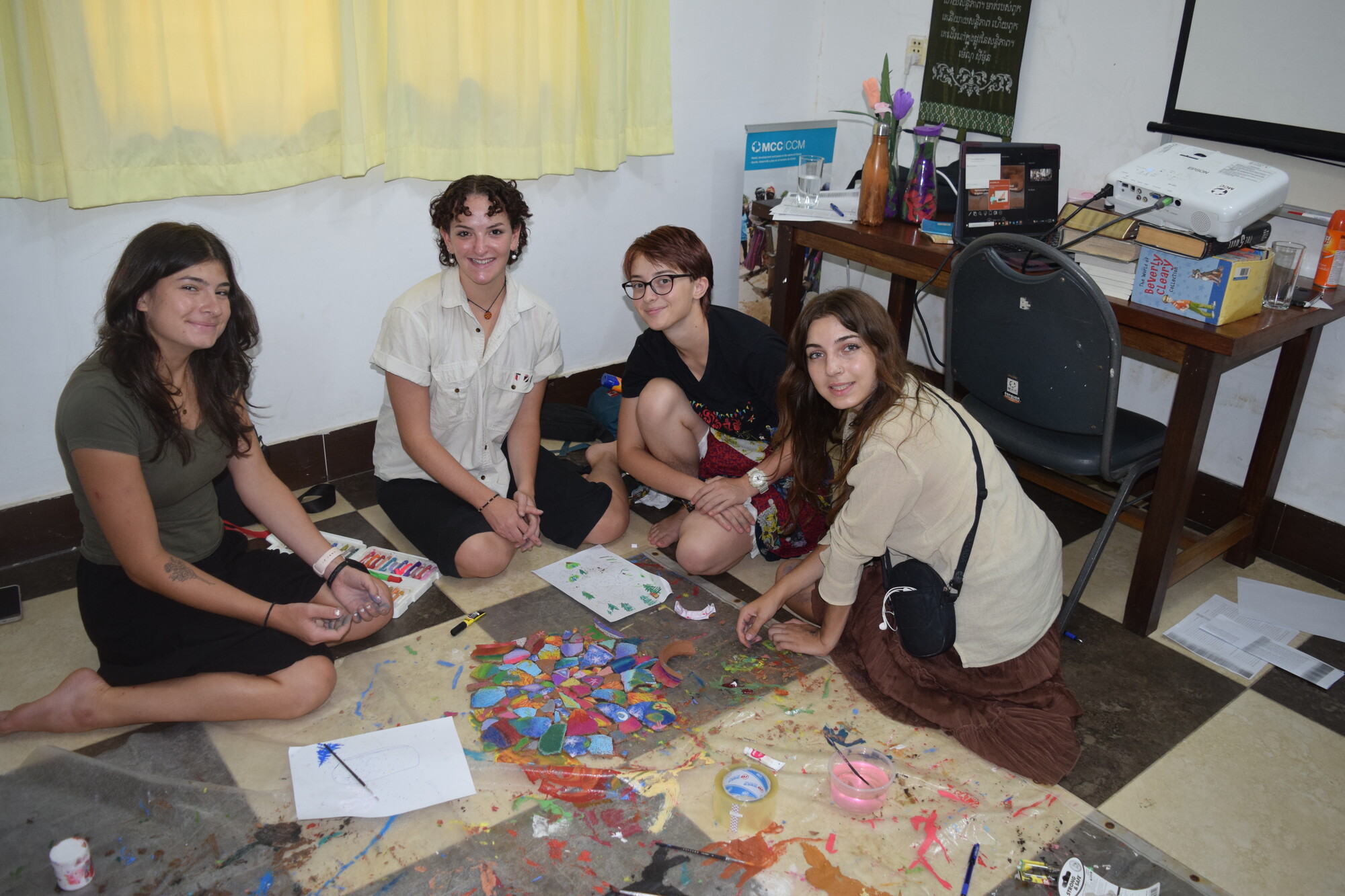 Four individuals are seated on the floor, surrounded by art supplies and colorful paper cutouts. They appear engaged in a creative activity, smiling at the camera in a room with a casual setting.