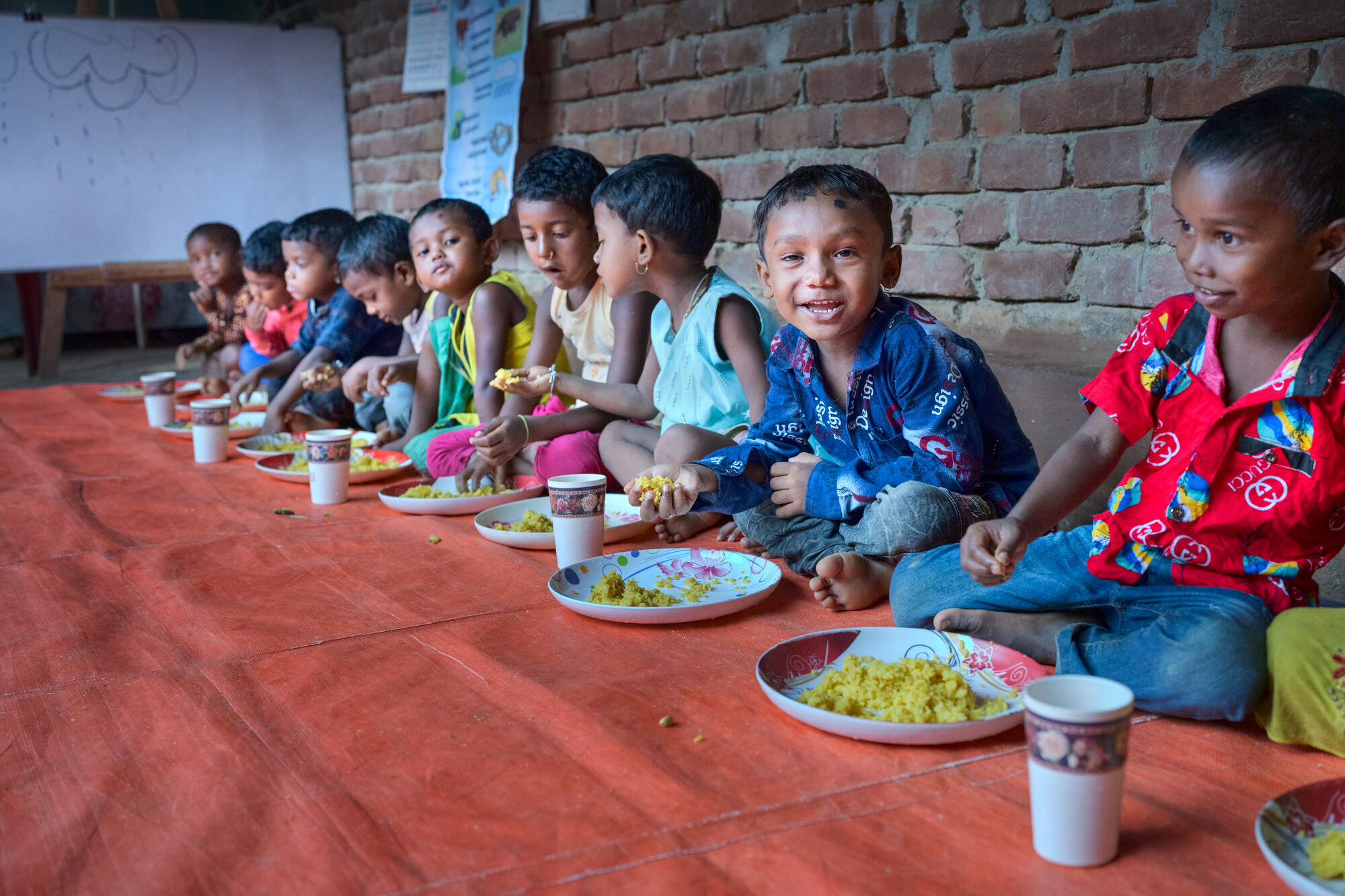 A group of children sit at one side of a table eating, with one smiling at the camera.