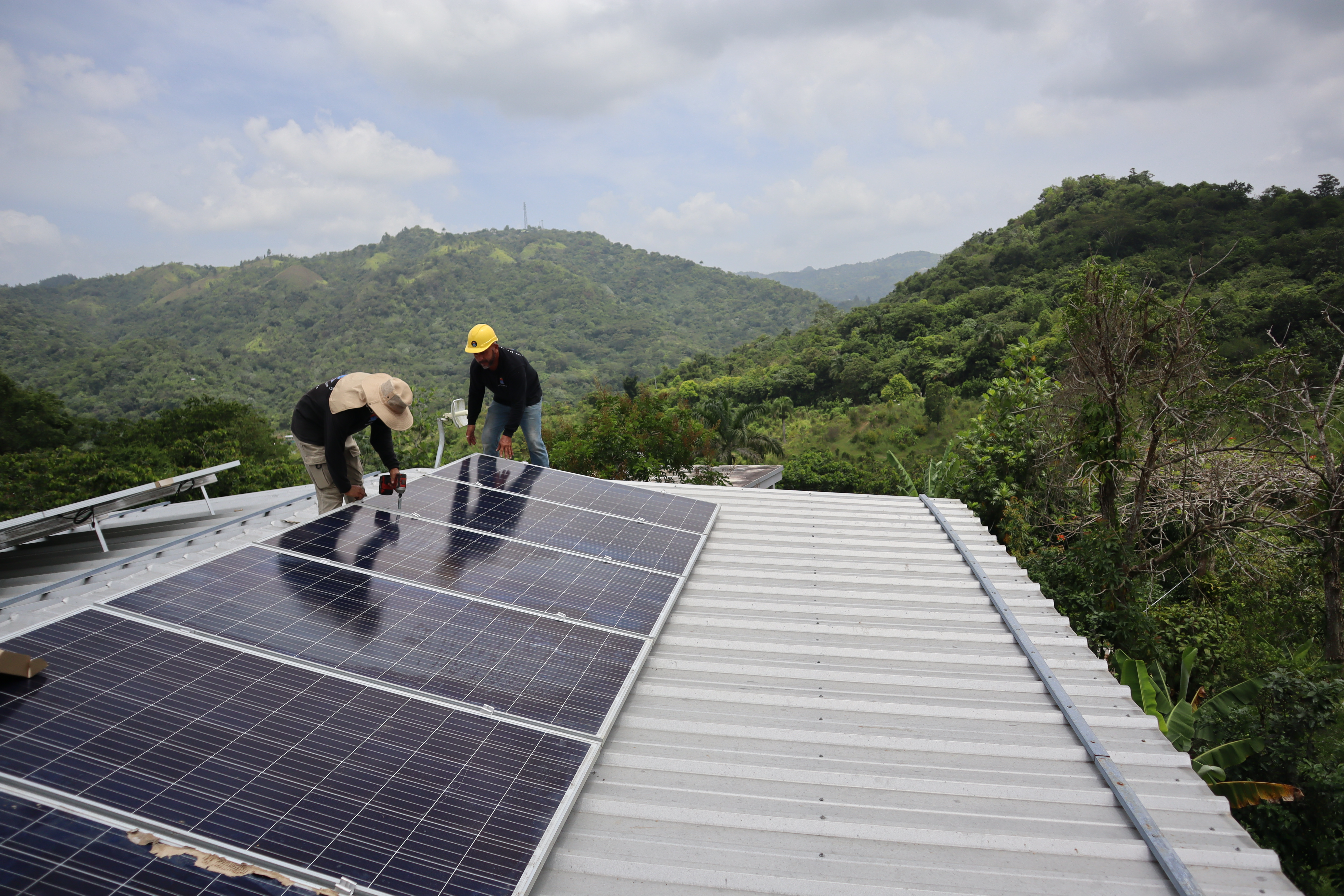 two workers install solar panels on a roof surrounded by mountain views