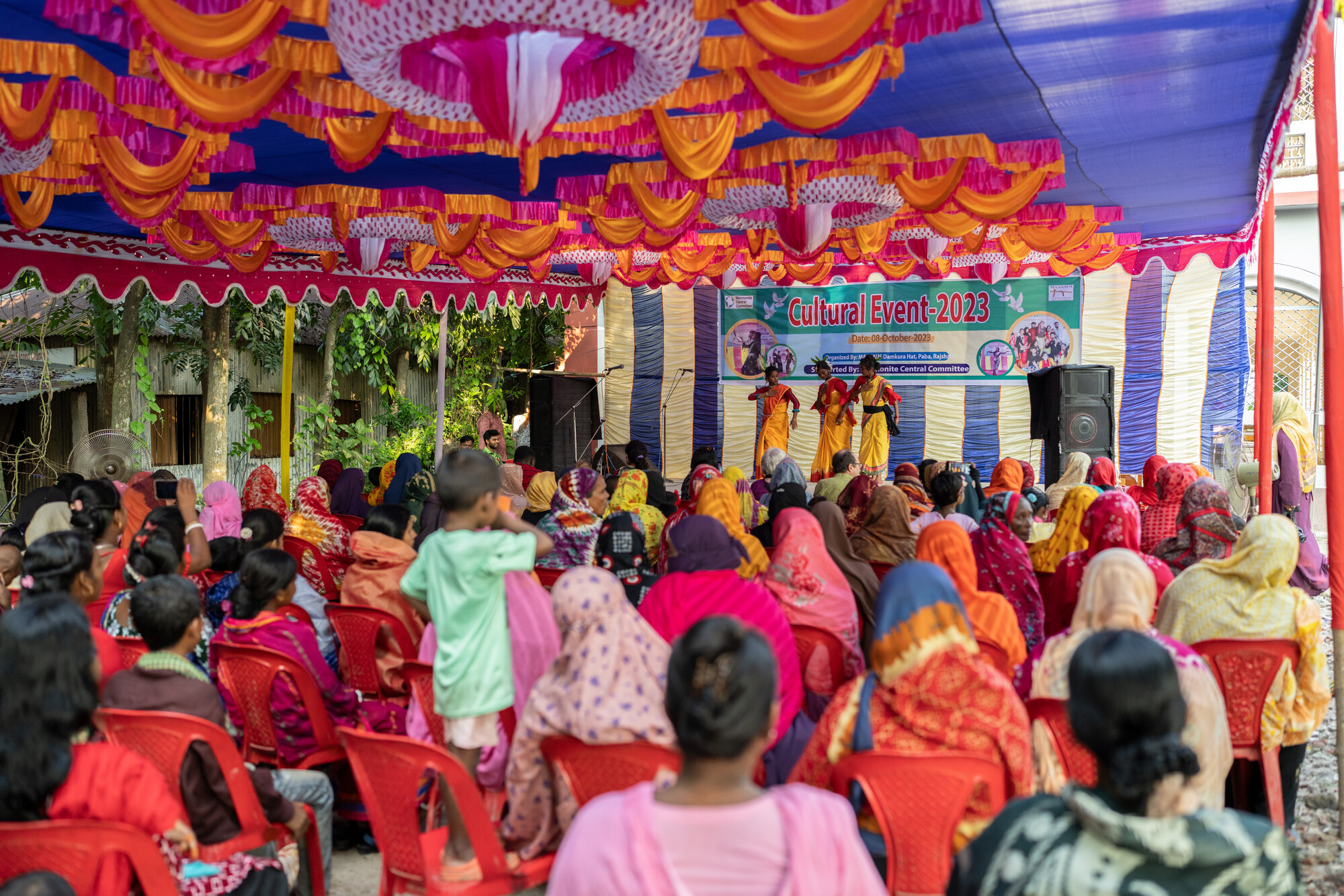 An audience in colorful attire watches a cultural event under a tent with performers on stage. Decorations hang overhead, and a banner reads "Culture Event 2023."