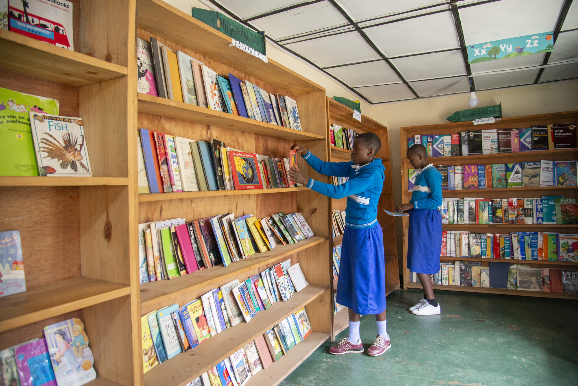 Two students wearing blue uniforms select books in a library.