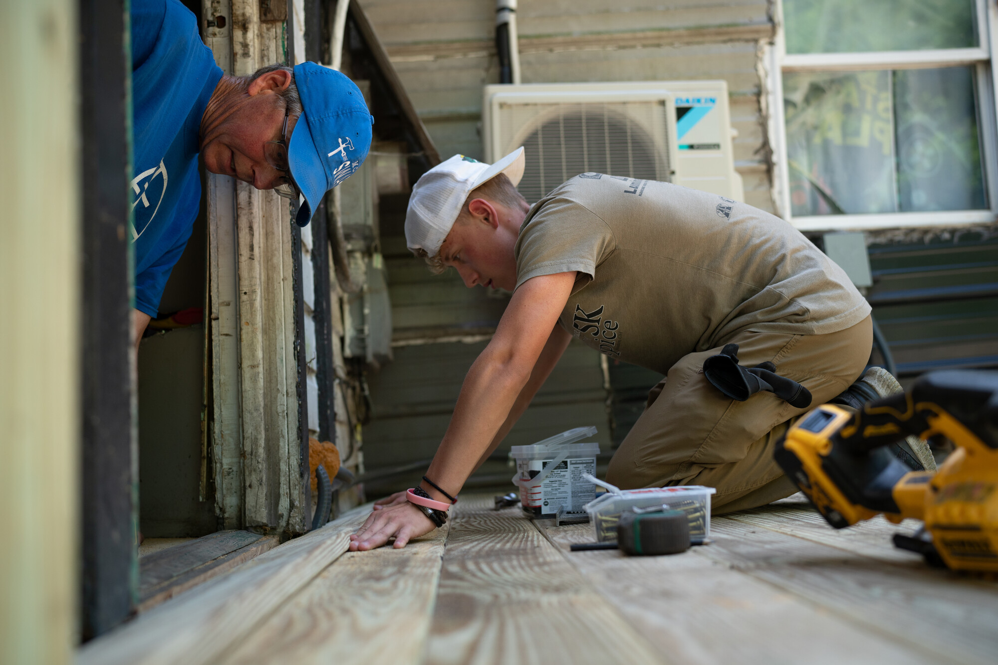 Two individuals are working on a wooden floor installation. They are using tools and appear focused on their task. The setting suggests a home renovation project.