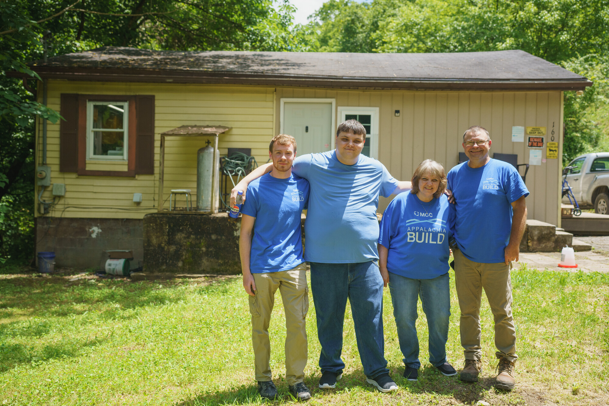 Four individuals in blue shirts stand smiling in front of a modest house with greenery around. It's a sunny day, and they seem to be in a residential area.