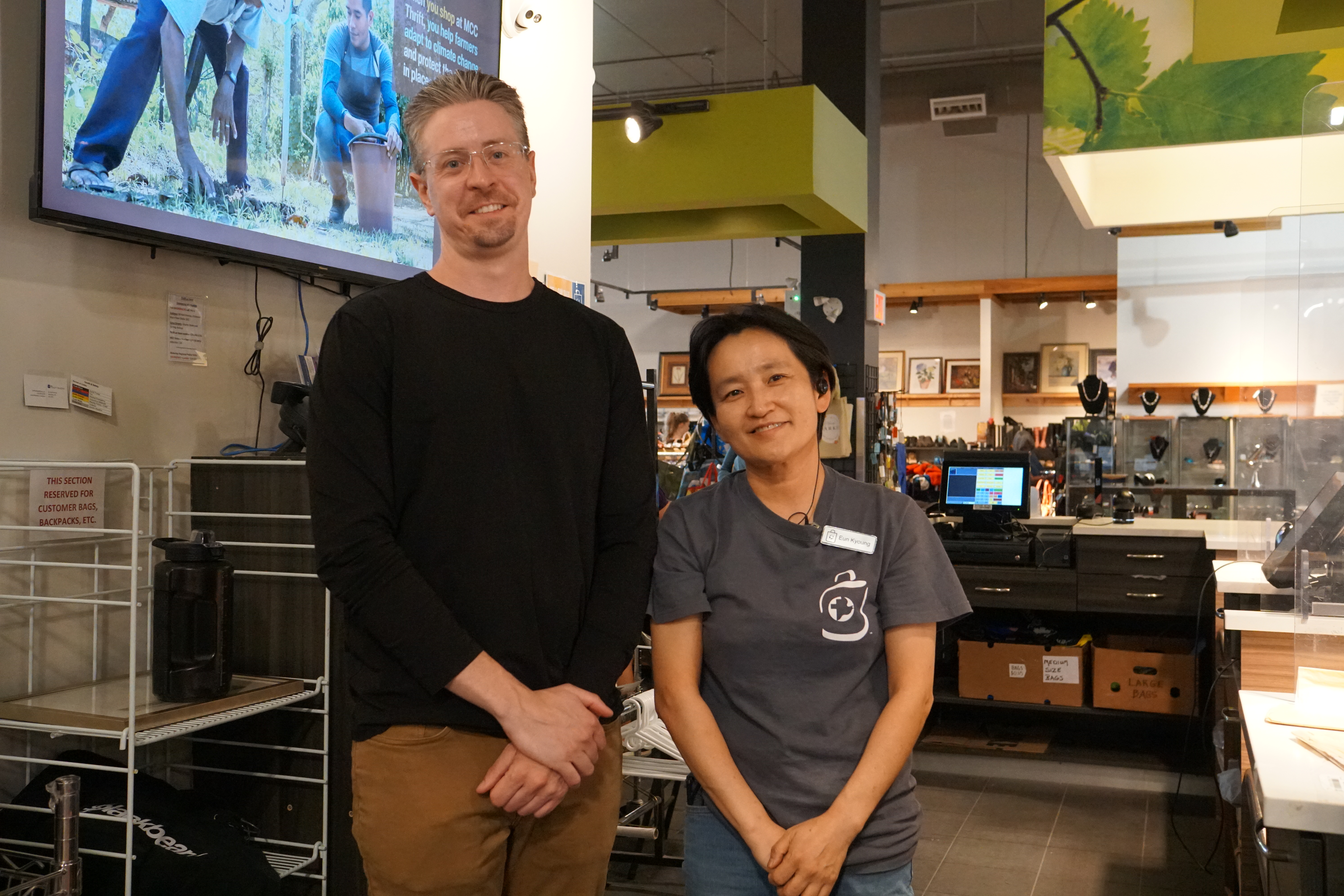 A man in black shirt stands next to a shorter woman in a grey t-shirt, they are behind the cash register of a retail clothing store.