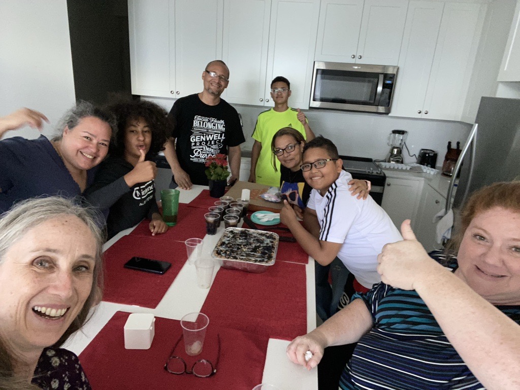 A group of people gather with big smiles around a kitchen table with a birthday cake on it.