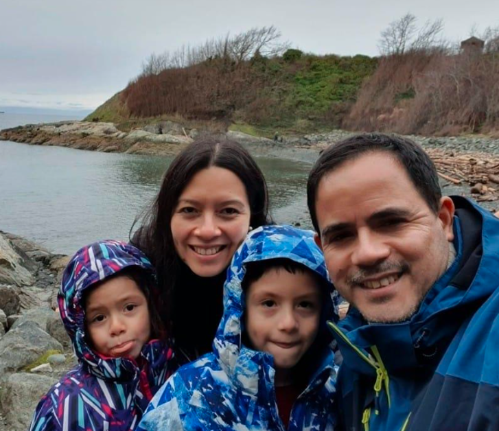 A picture of a family of four: a mother, father, son and daughter in front of a body of water and trees in the backgorund