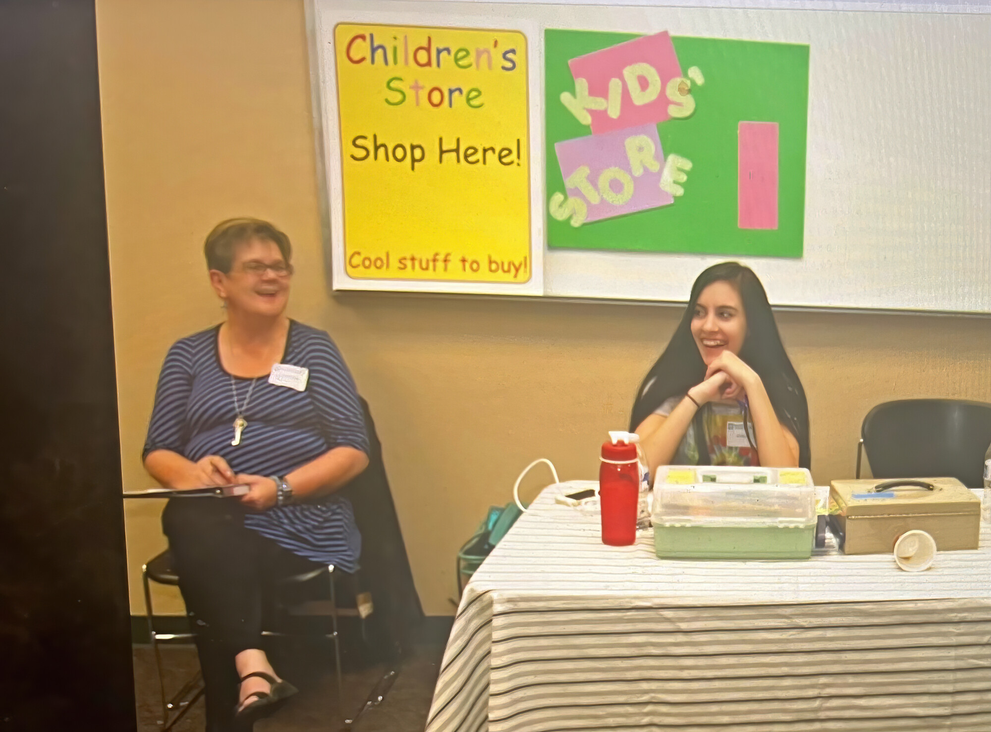 The photograph features a woman and her teenaged granddaughter seated at a table in a room. Behind them, there are signs promoting a children's store and a display of items on the table.