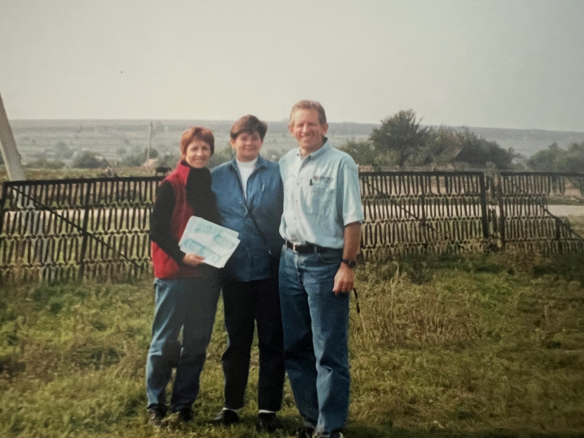 Wearing long-sleeved shirts and jeans, Rick Martens (right) stands with his wife, Carole Martens (center), and sister, Catheryn Martens (left), in a grassy field that was once a family farmstead.  The Martens family visited the land, located in what is now Dolynske, Ukraine, in 2002.