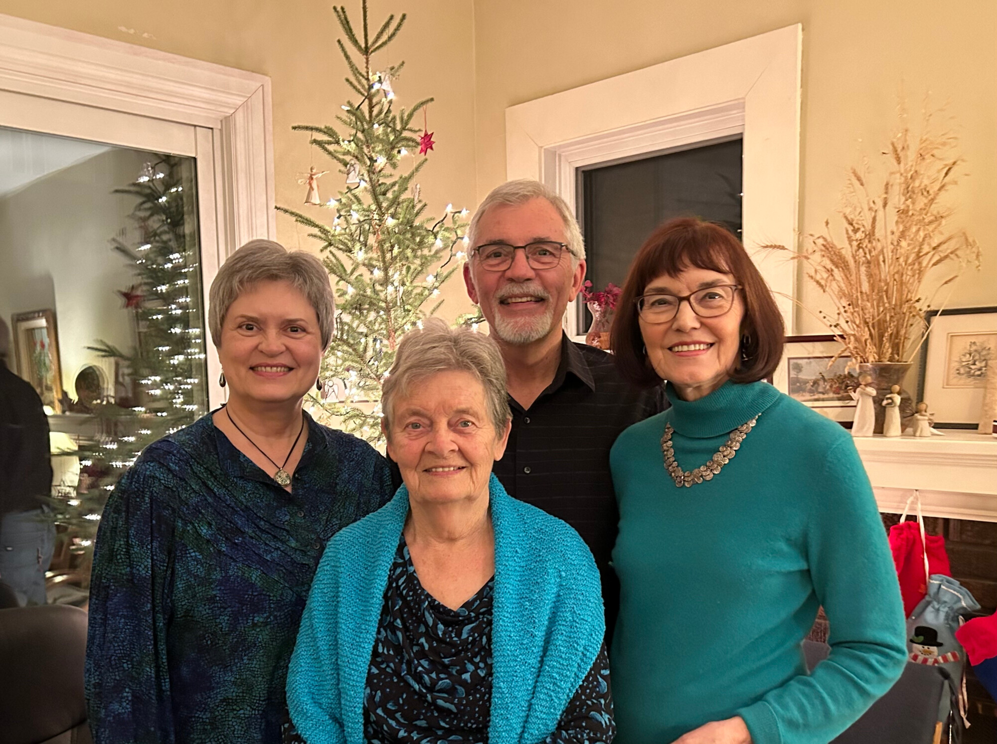  The photograph features four individuals standing together in a cozy indoor setting, with a decorated tree in the background. They are smiling and appear to be enjoying a festive gathering, showcasing a warm and familial atmosphere.