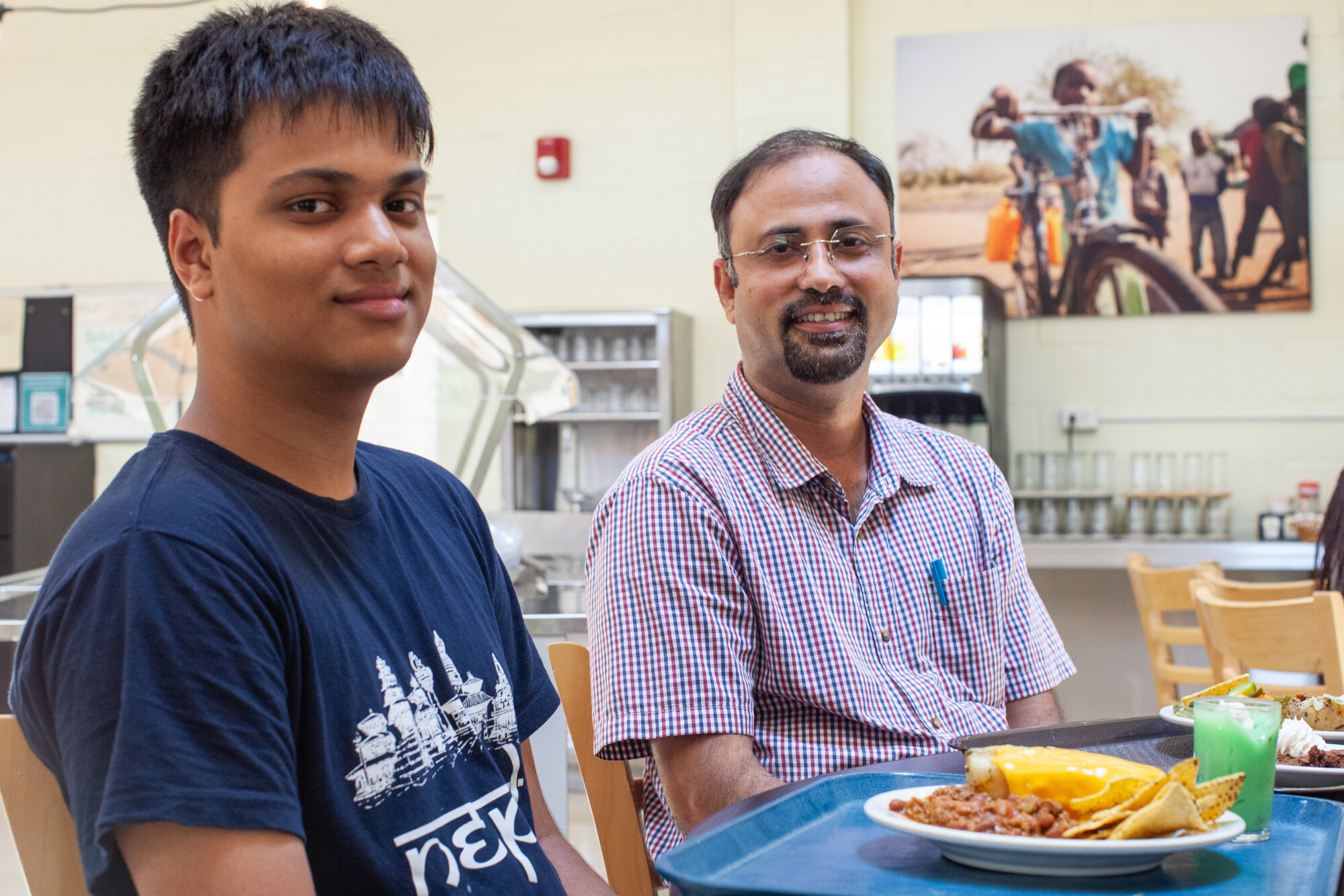 Two people sitting next to each other with food in front of them in a cafeteria look at the camera.