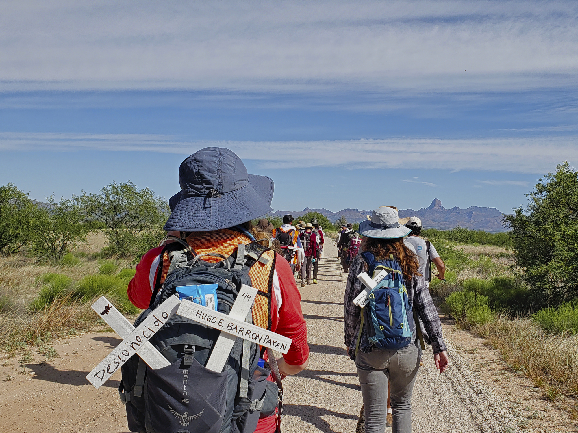 A group of people are walking on a dirt trail. There are crosses in their backpacks with messages in Spanish.