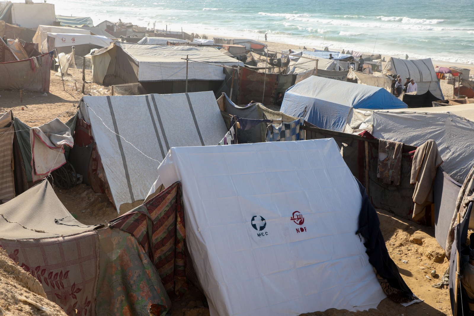 The photograph shows a cluster of tents in a sandy area near a beach, with some people visible in the background.