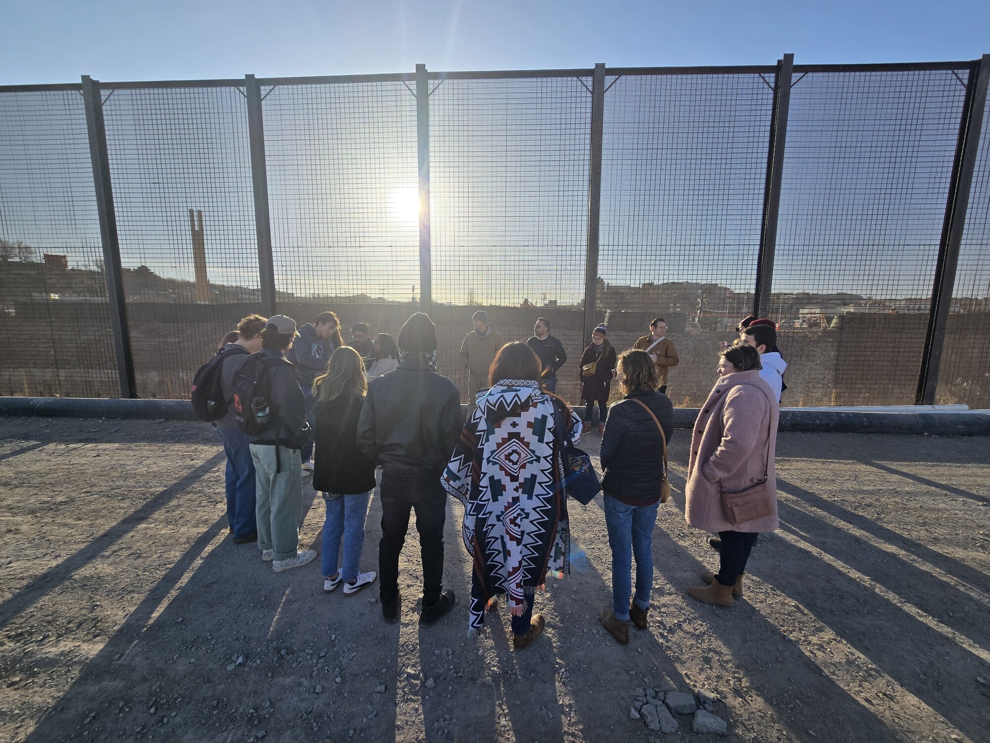 group of people circled in front of border wall between the US and Mexico