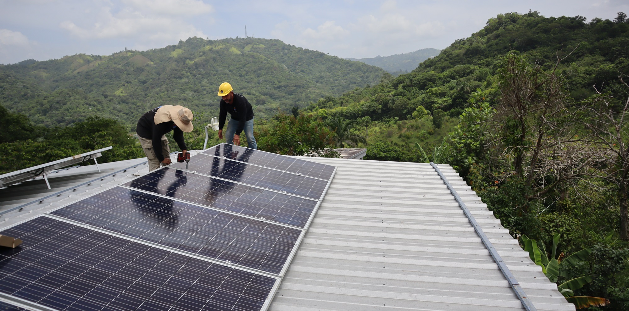 Two men install solar panels on a roof with mountains in background.