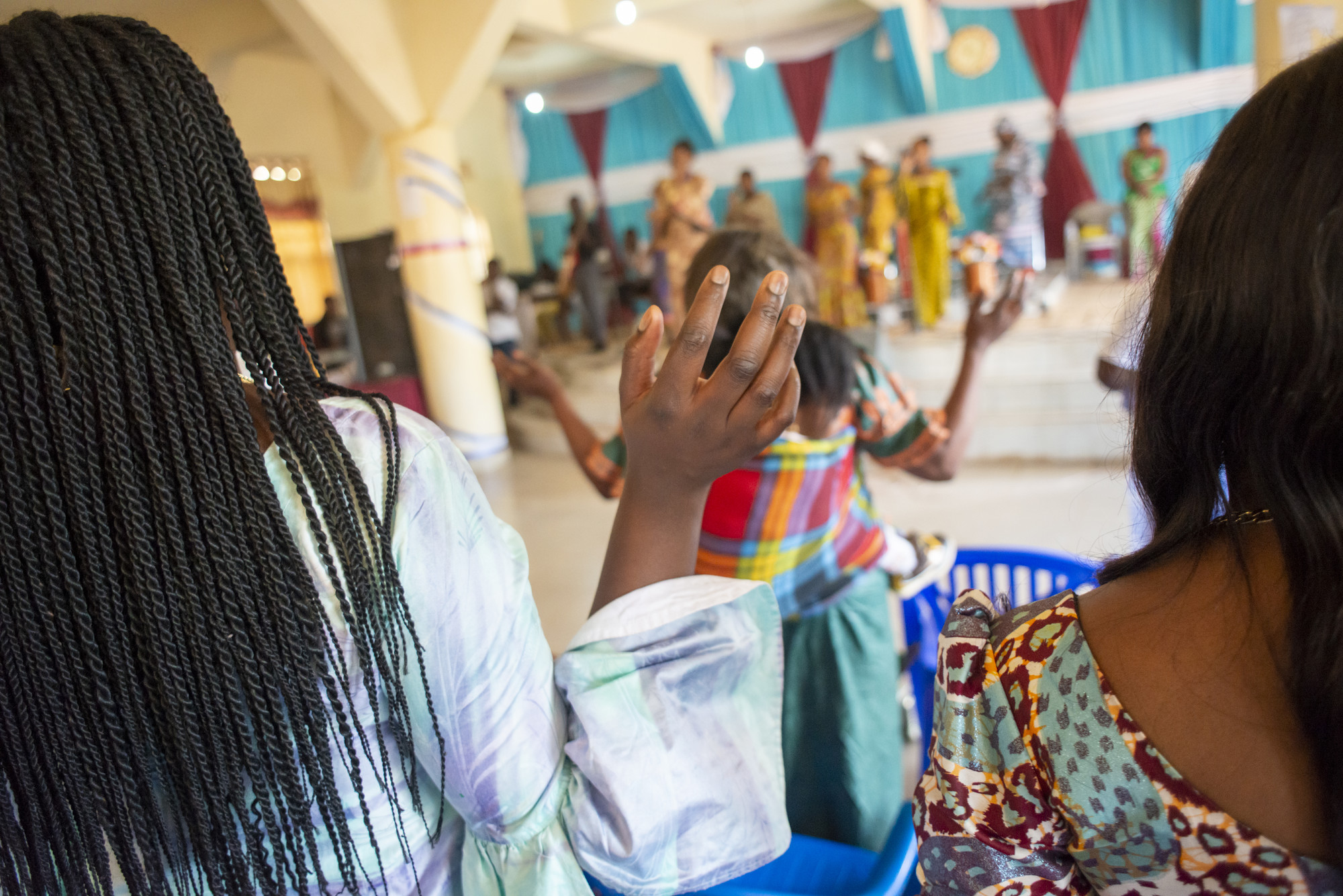 Two women with back to camera raise hands while watching women dance in front of church