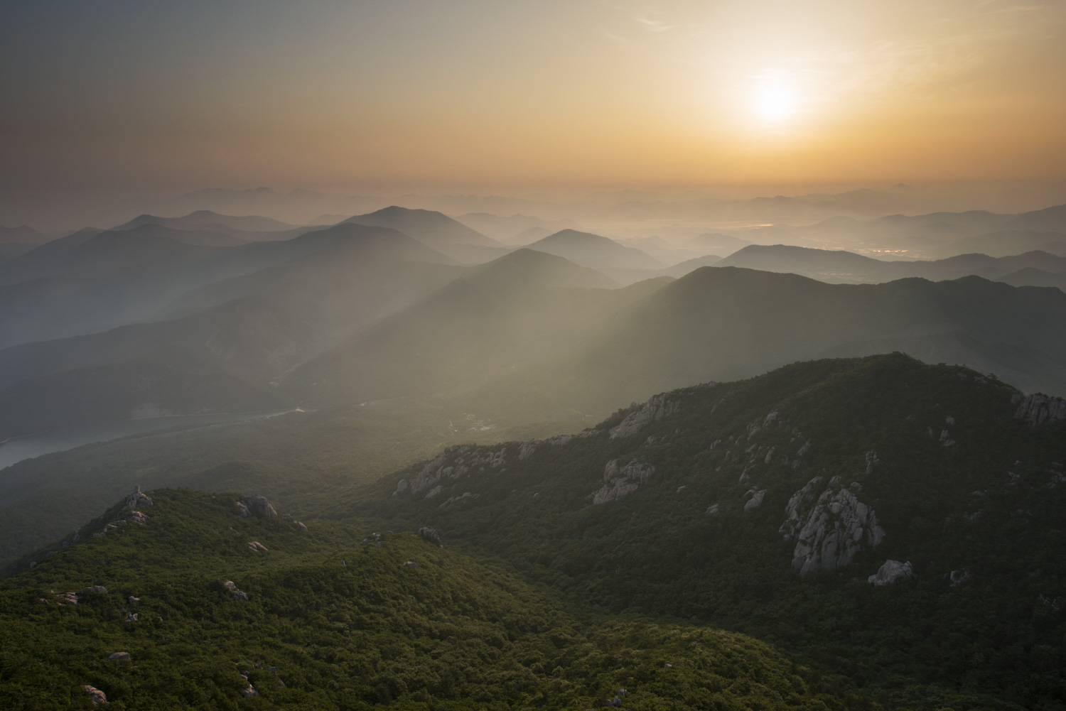 A photo of dawn over mountains in Korea, there is a layer of fog at the mountaintops and soft orange and yellow light as the sun peeks through.