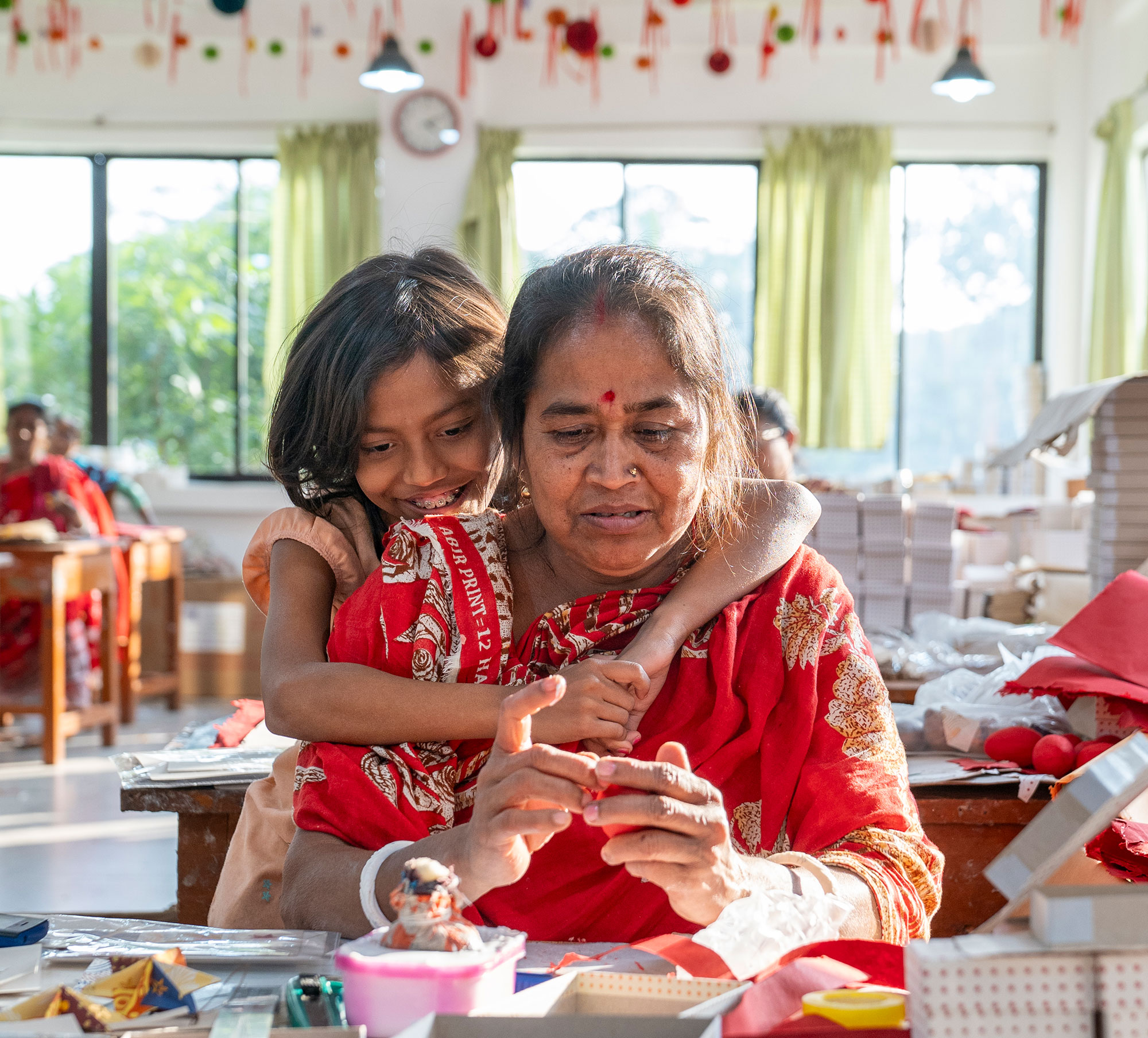 A woman sits working at a table while a young person embraces them from behind.