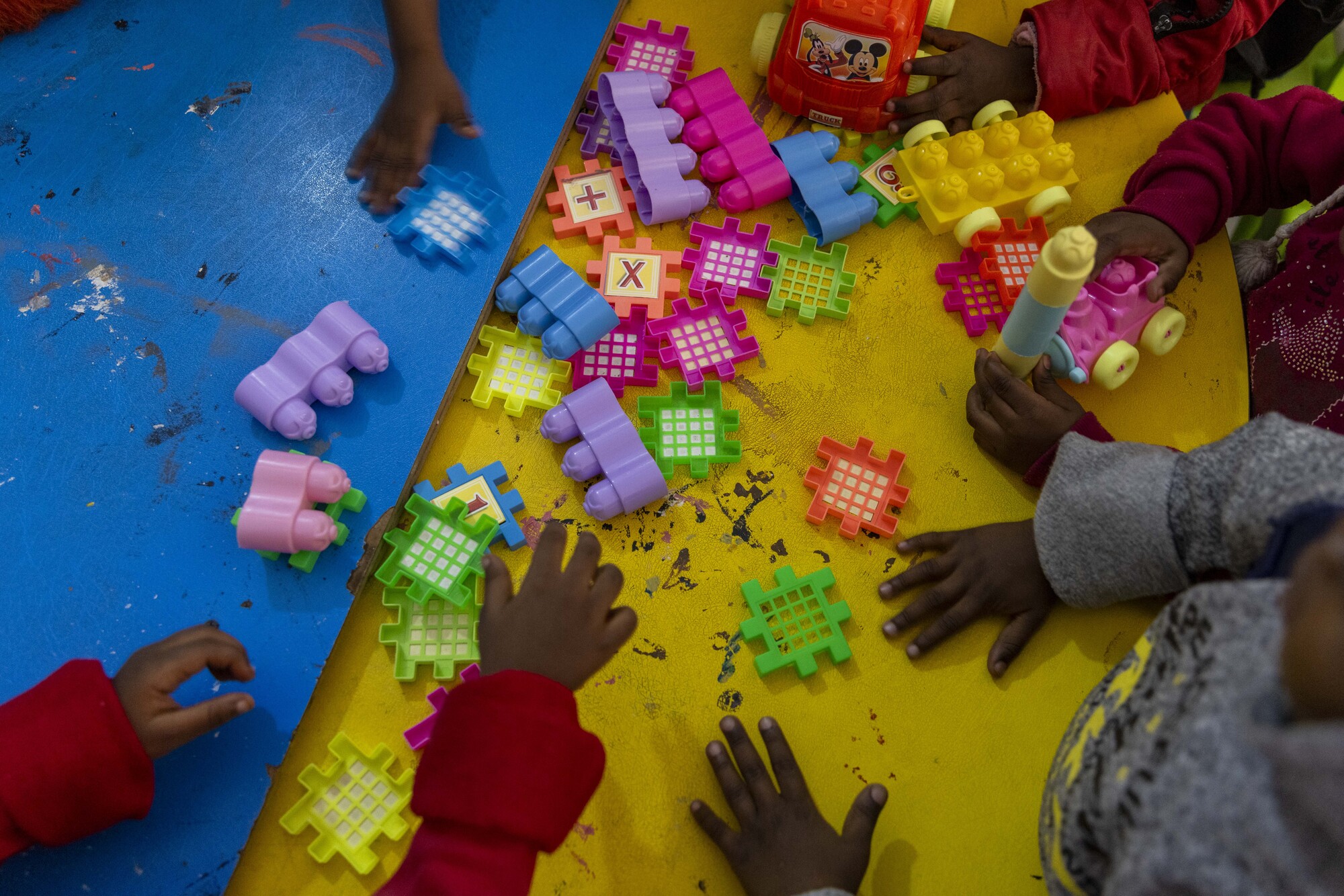 The image shows hands of children playing with various building blocks and toys scattered across two colorful tables.