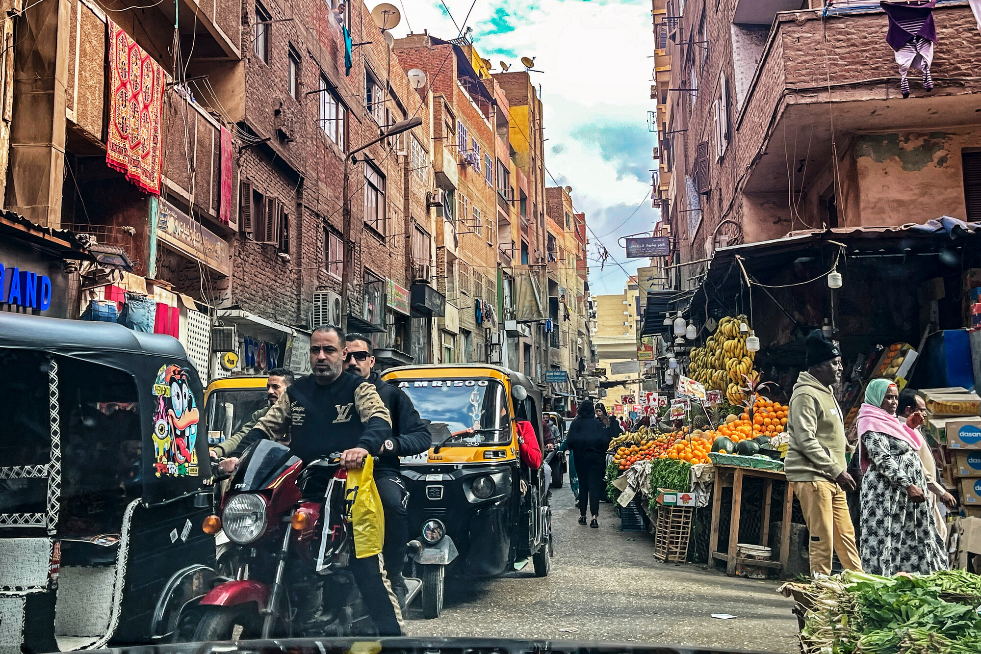 A busy street scene with vendors, fruit stalls, and vehicles. People walk alongside motorcycles and auto rickshaws in a narrow alley.