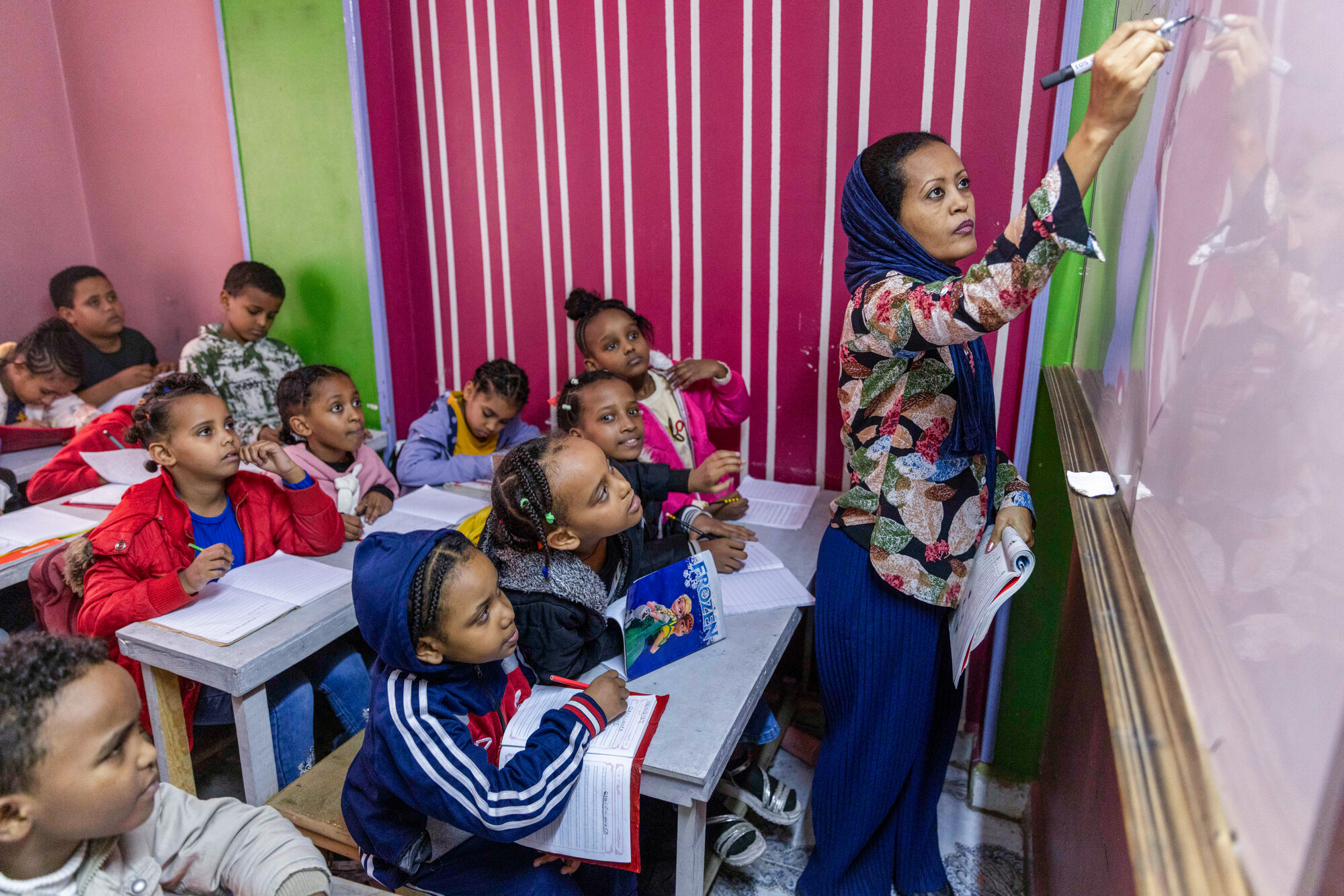 A teacher writes on a board while students attentively watch. Desks are arranged in rows, and the classroom has colorful walls.
