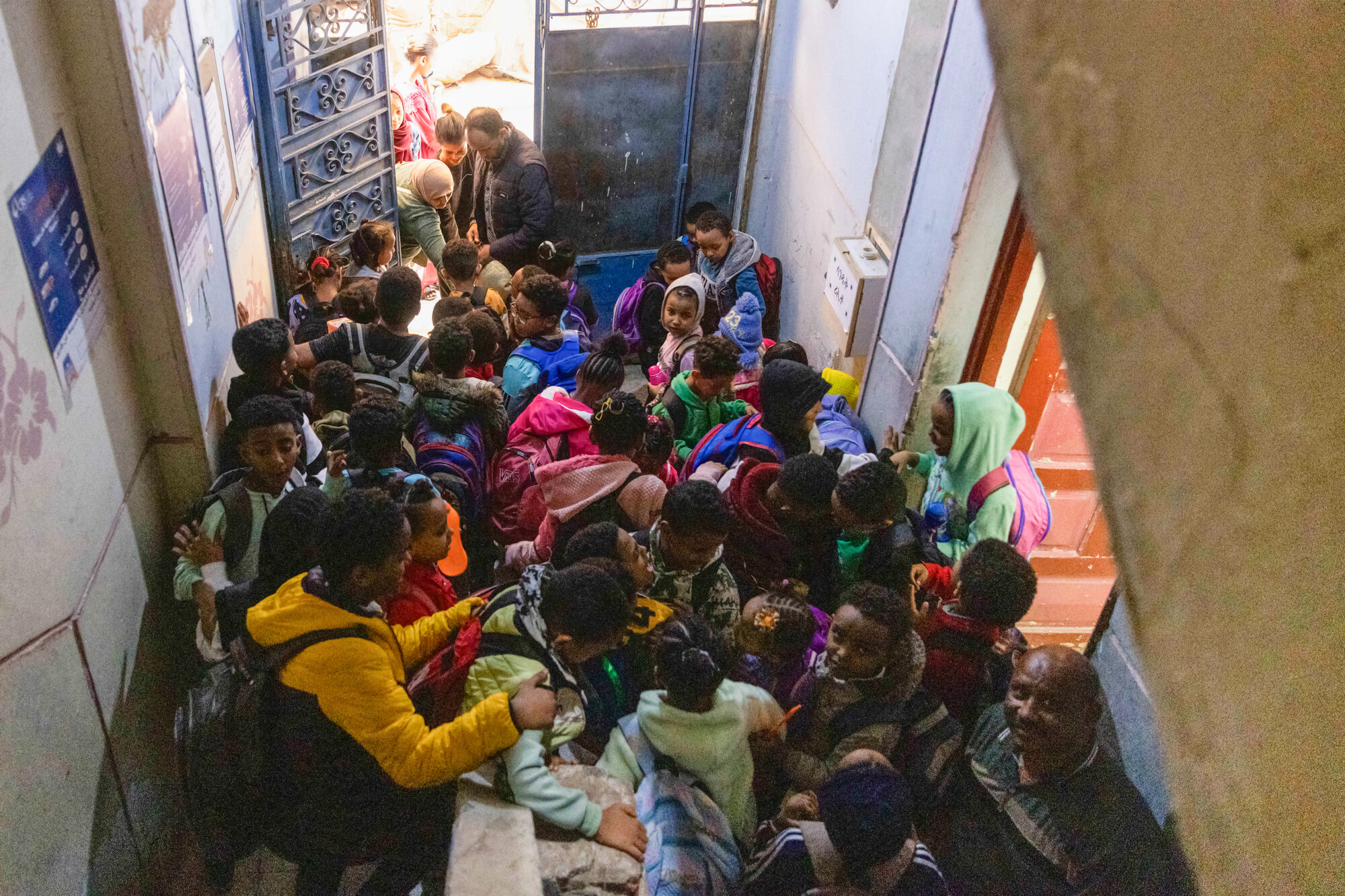 A crowded staircase filled with children and adults, some carrying backpacks, in a narrow indoor space.