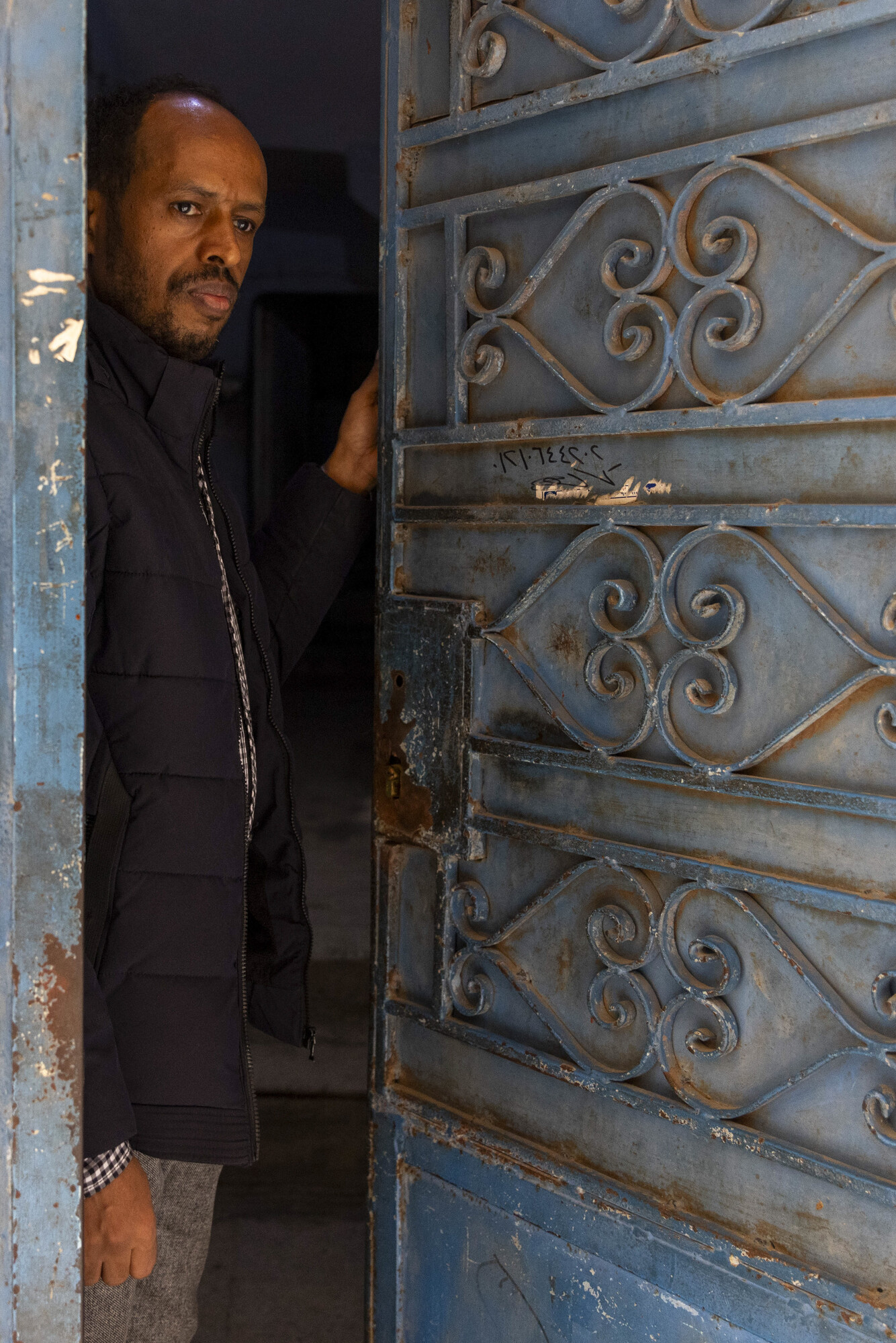 + A man stands at an ornate door, partially open, with intricate patterns. He gazes thoughtfully, framed by the doorway.