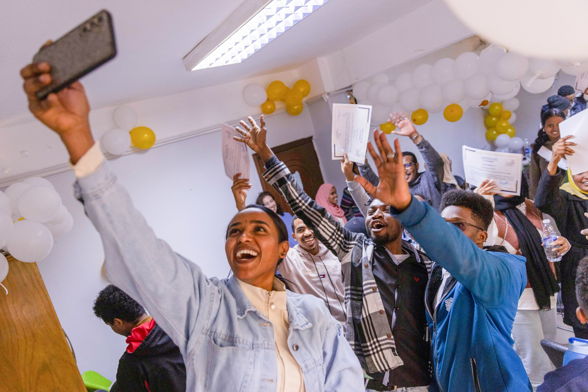 A group of people are celebrating indoors, holding certificates and taking selfies, with balloons in the background.