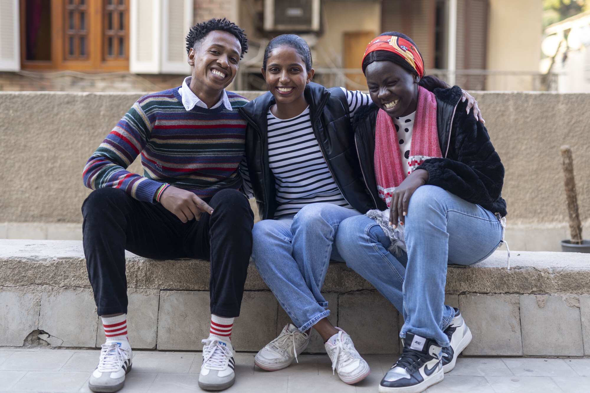 Three individuals sit closely on a stone bench, smiling and posing together. They wear casual clothing and appear relaxed.