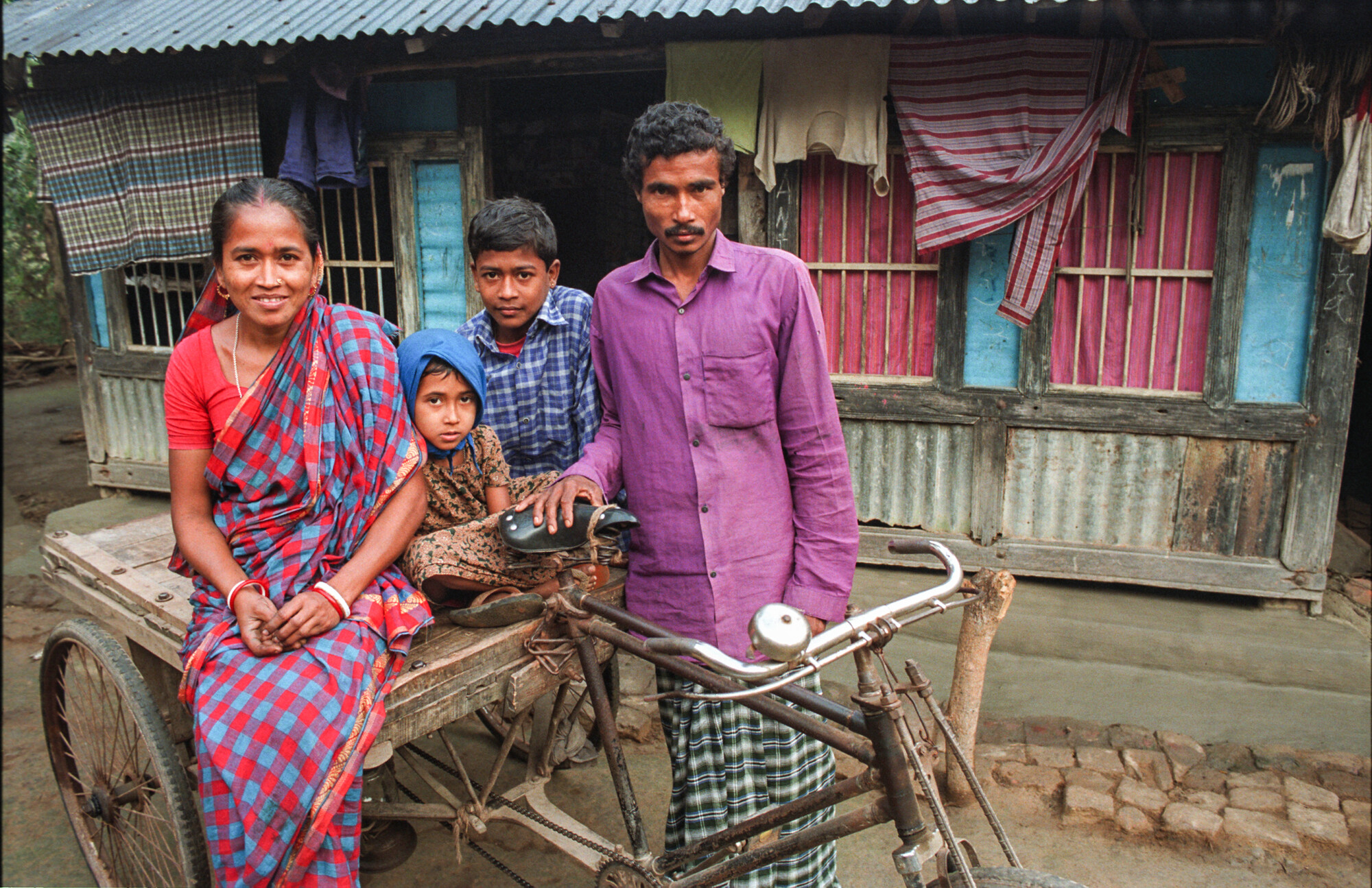 A woman, man and two children pose on a cart in front of a wooden house with barred windows and hanging cloth.