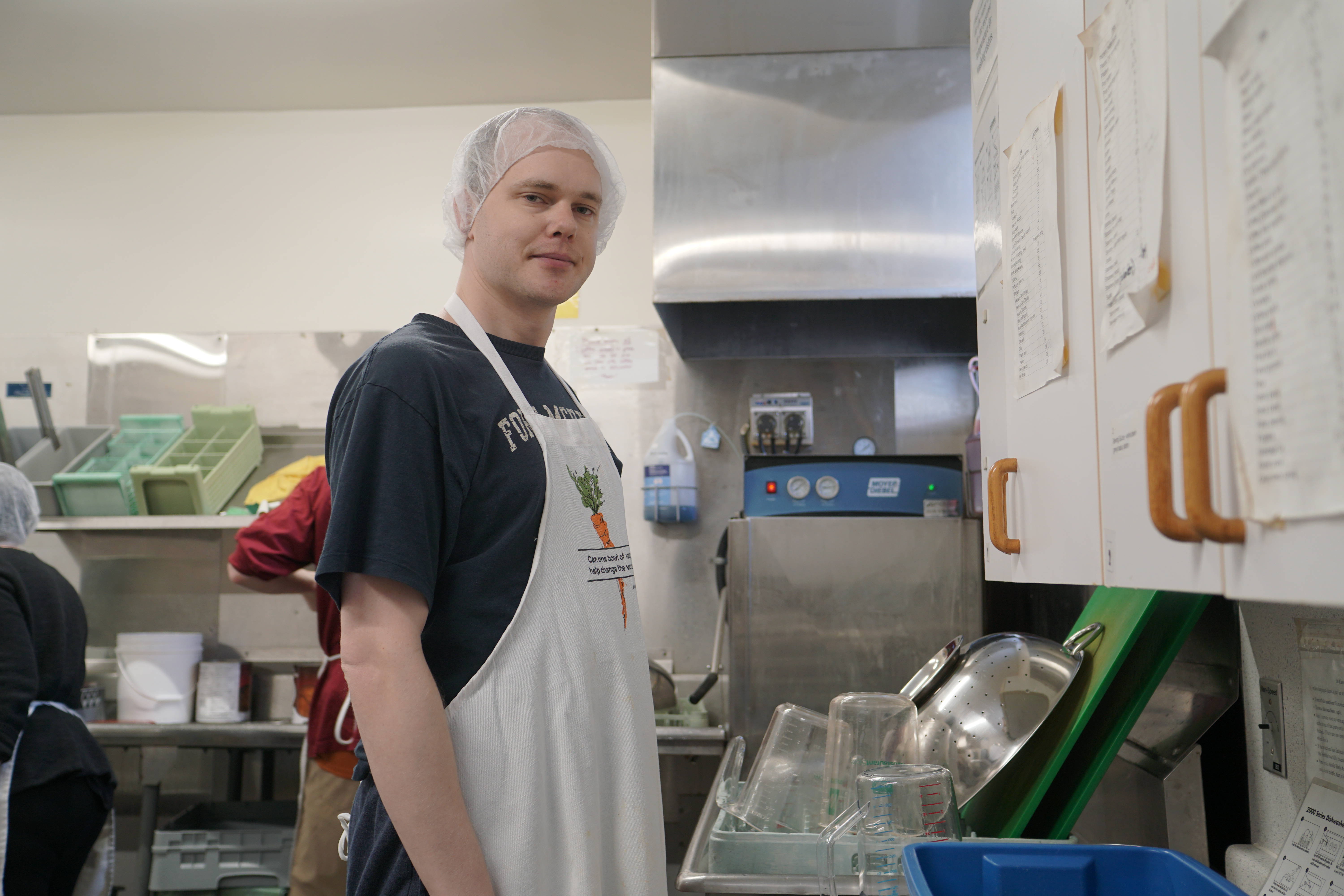 A man wearing a hair net and apron stands in an industrial kitchen