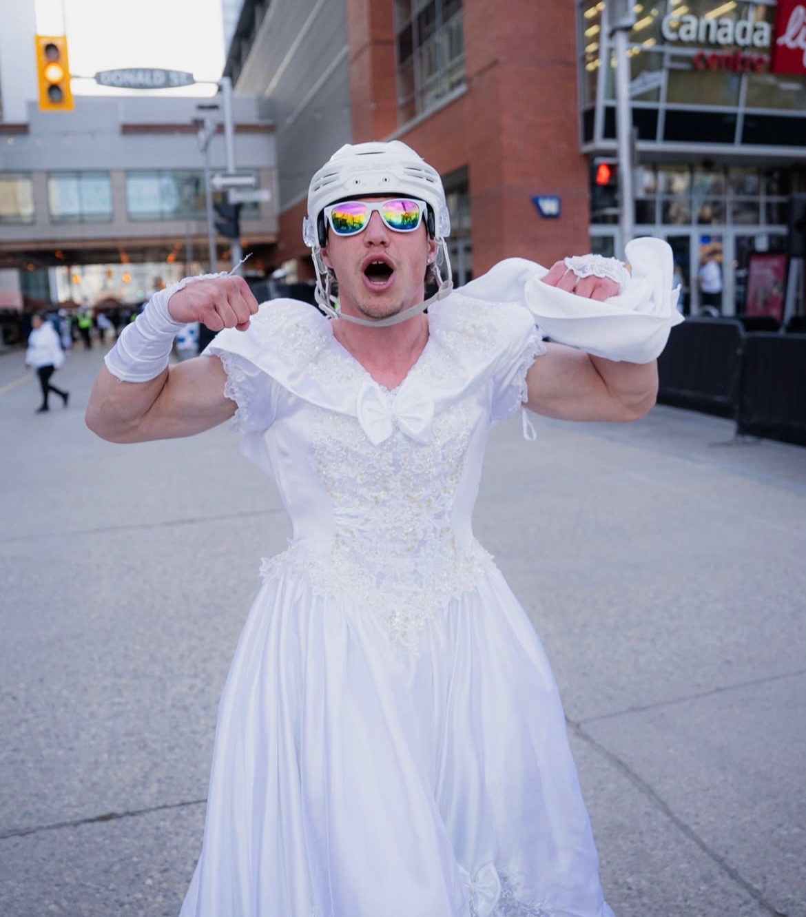 Man standing in downtown Winnipeg dressed in a white wedding dress, hockey helmet and sunglasses.