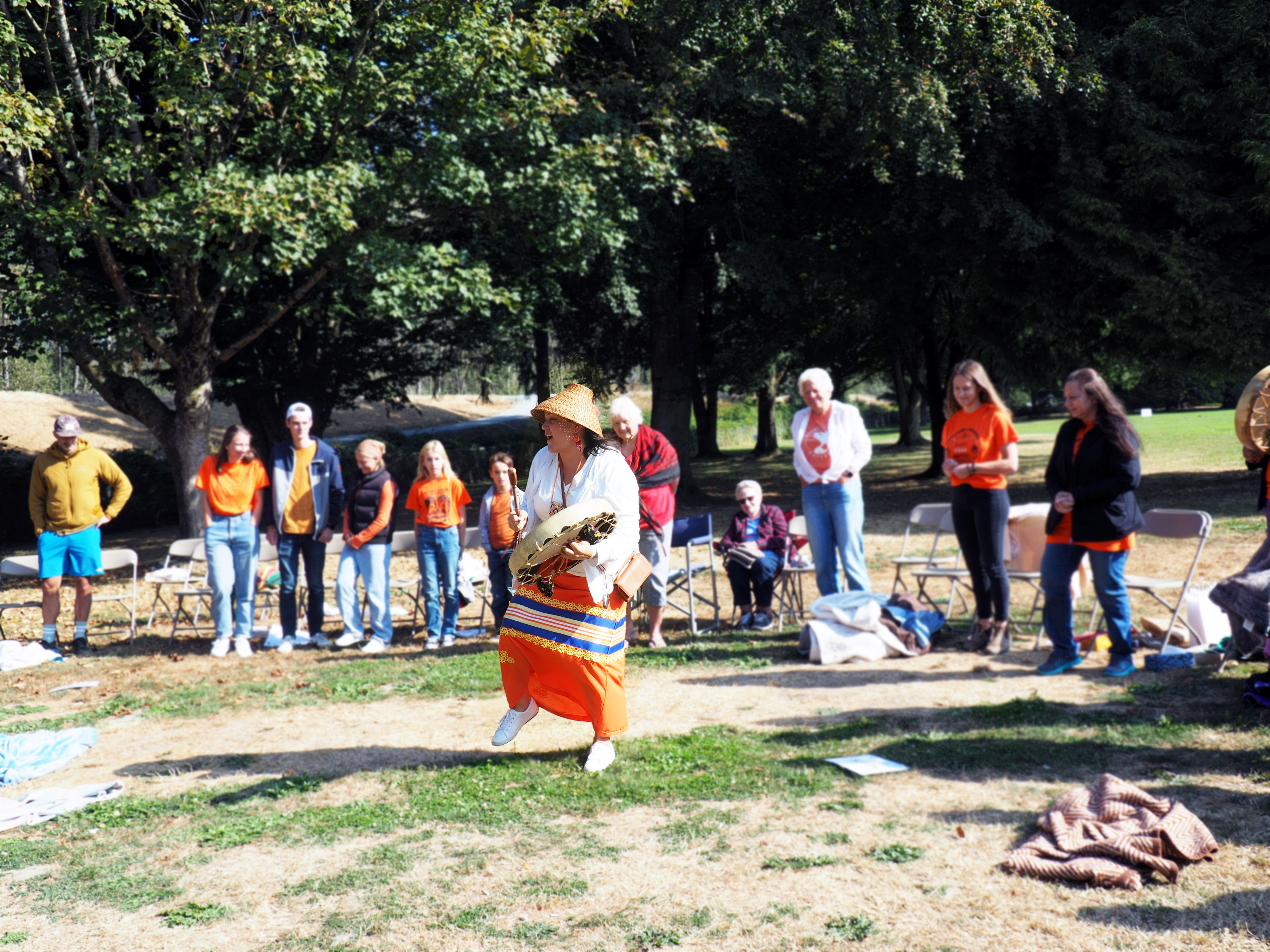 A person in traditional Indigenous regalia dances with a drum in front of a group of people during an outdoor cultural gathering.