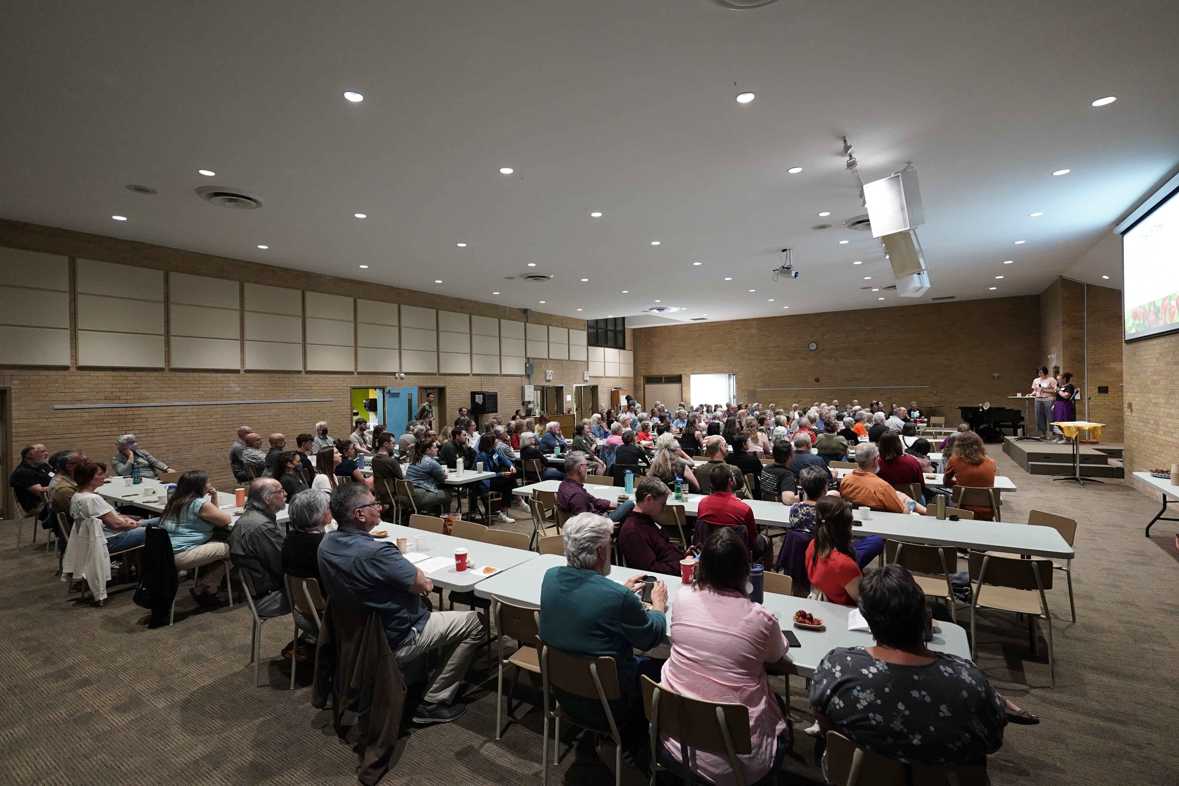 A crowd of people sit at long tables facing a stage where two people are speaking.