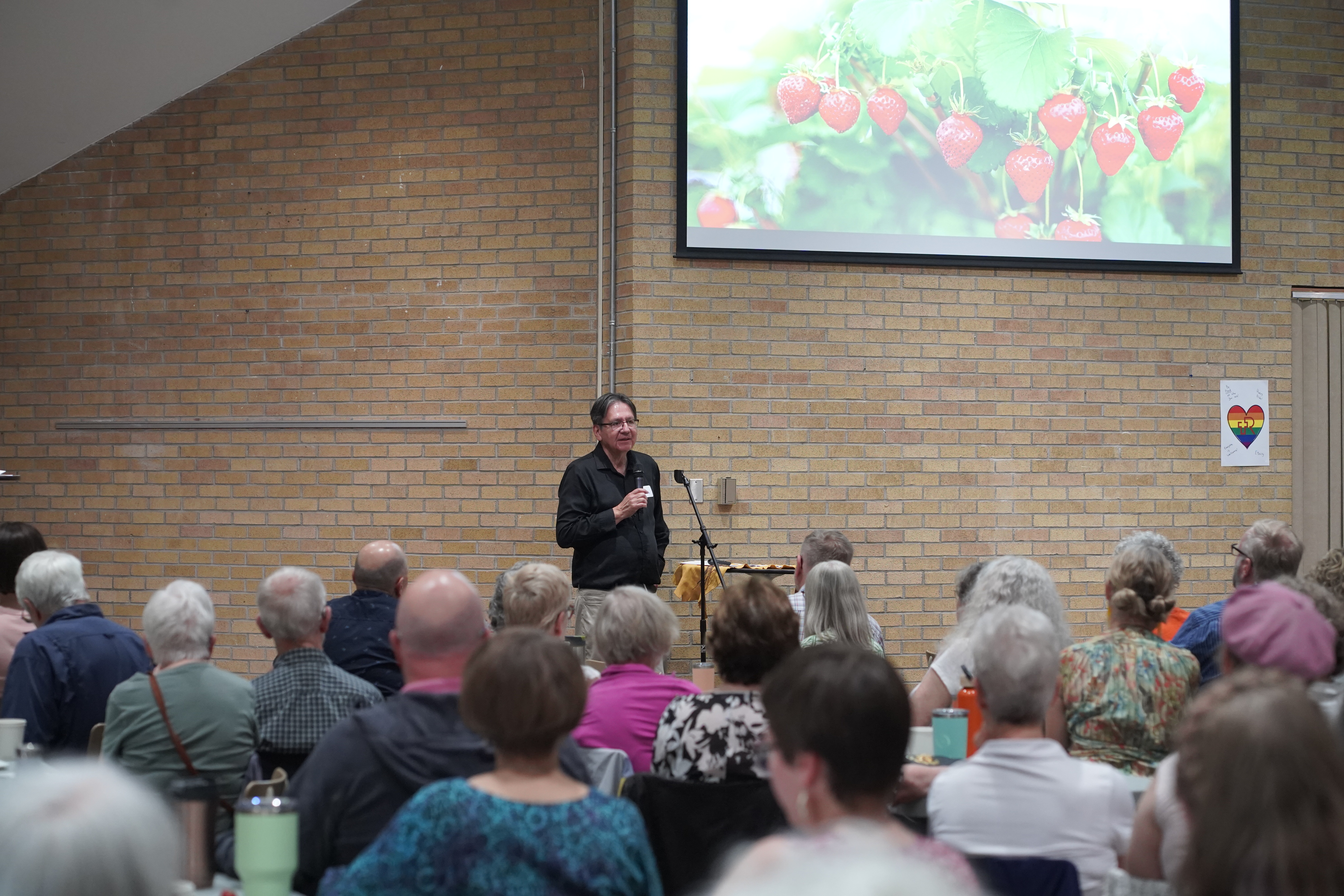 A man in a black shirt speaks to a large crowd with a slide behind him with a picture of strawberries on it
