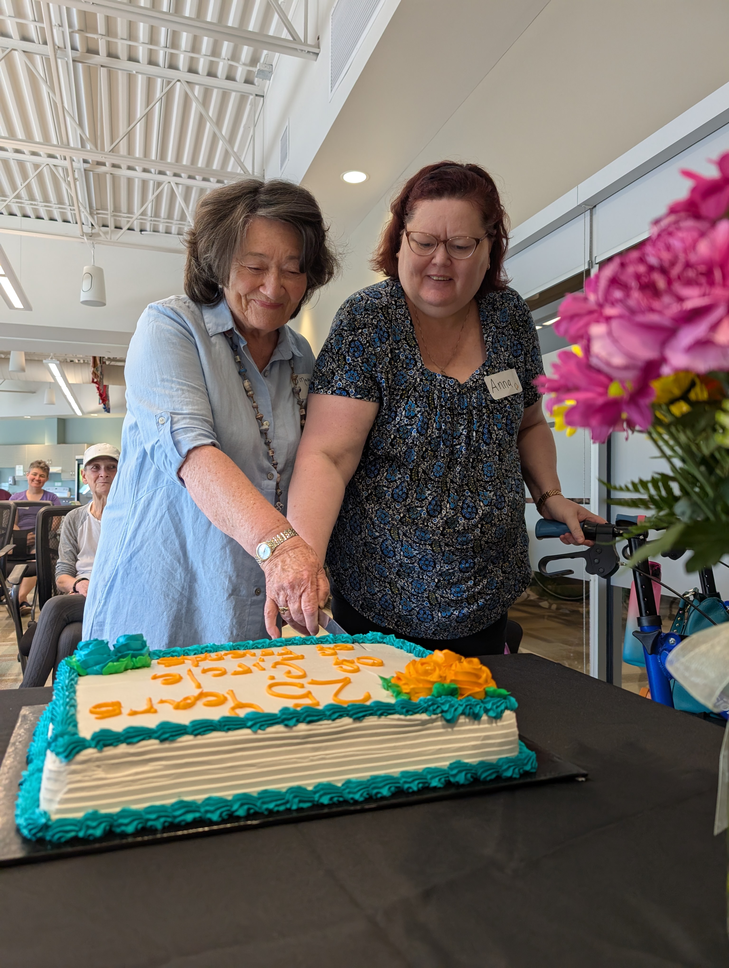 Two smiling women hold a single knife as they cut a decorated cake.