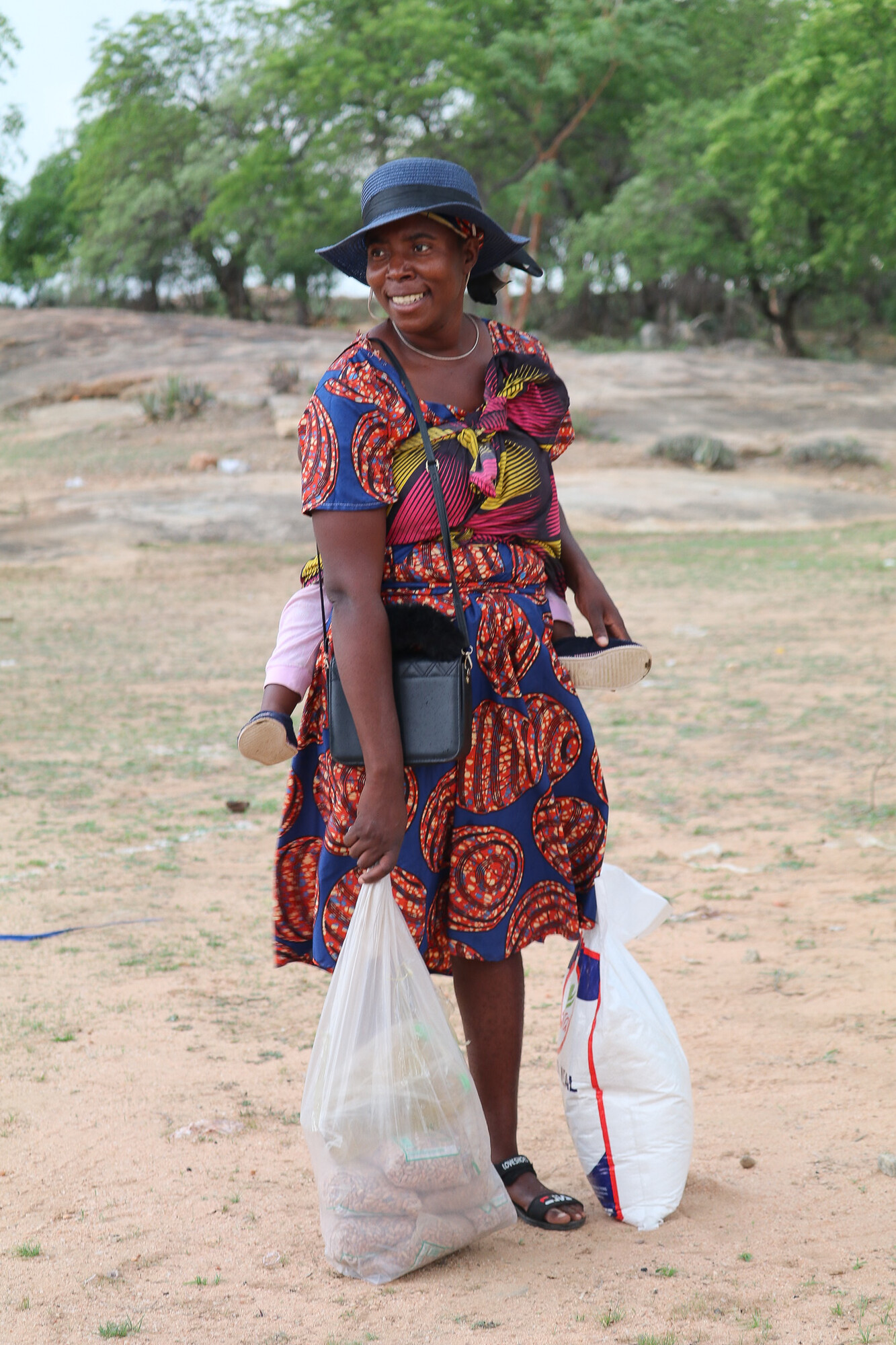 woman holding baby on back with bags of ground corn and beans at her feet