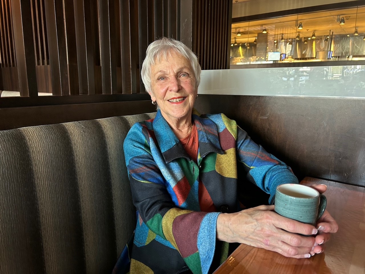 Legacy donor, JoAnne Janzen, sits in a booth with mug in hand. 