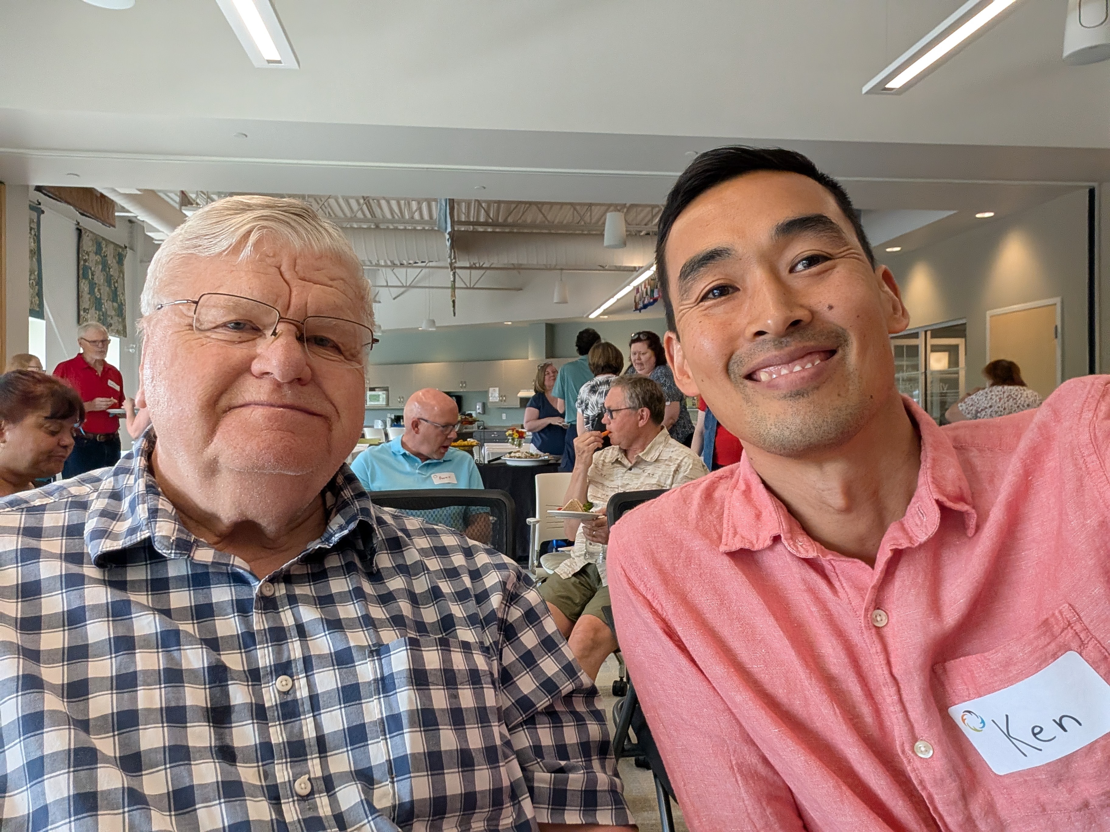An older white-haired man and a young Asian man in a pink shirt take a selfie together.