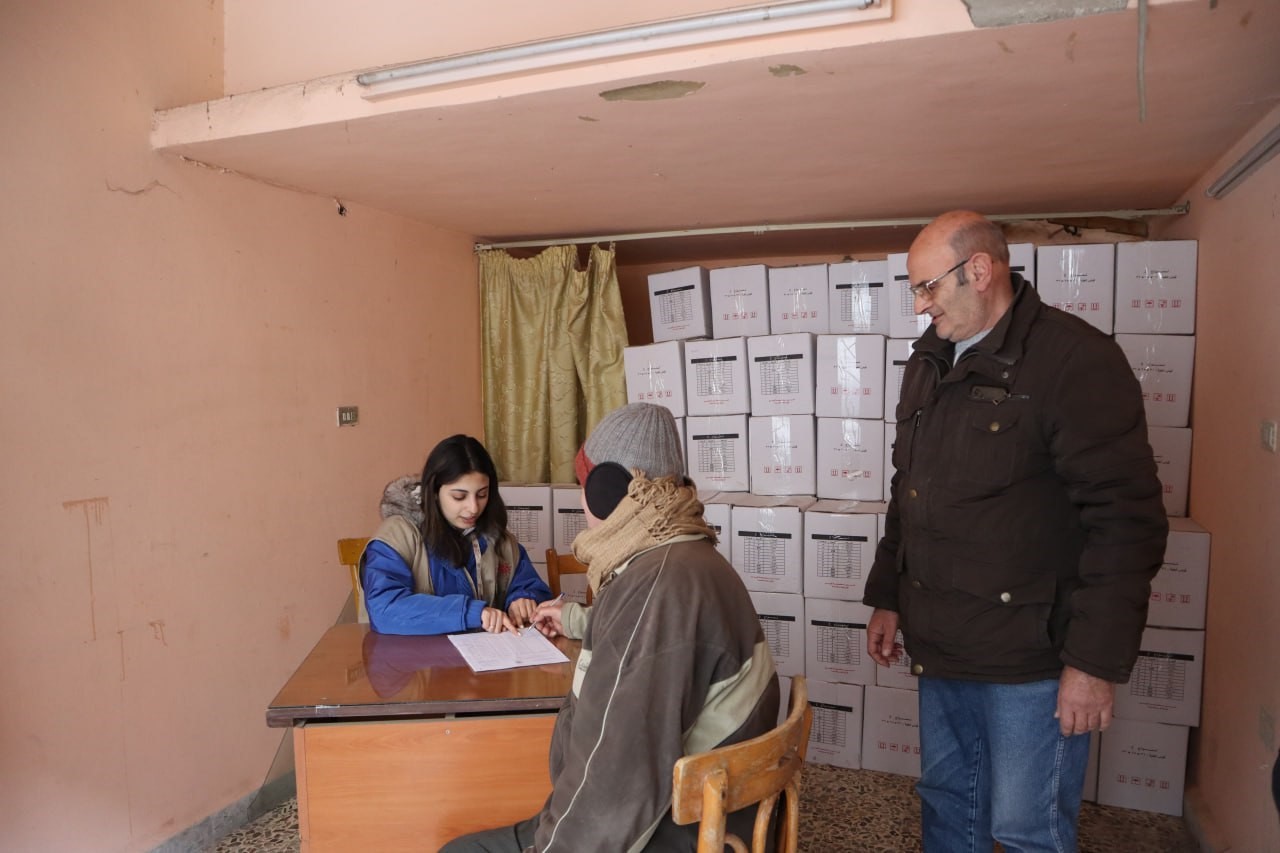 A woman sitting at a desk speaks to a person sitting in a chair. A man stands behind them next to a stack of boxes with relief food supplies.