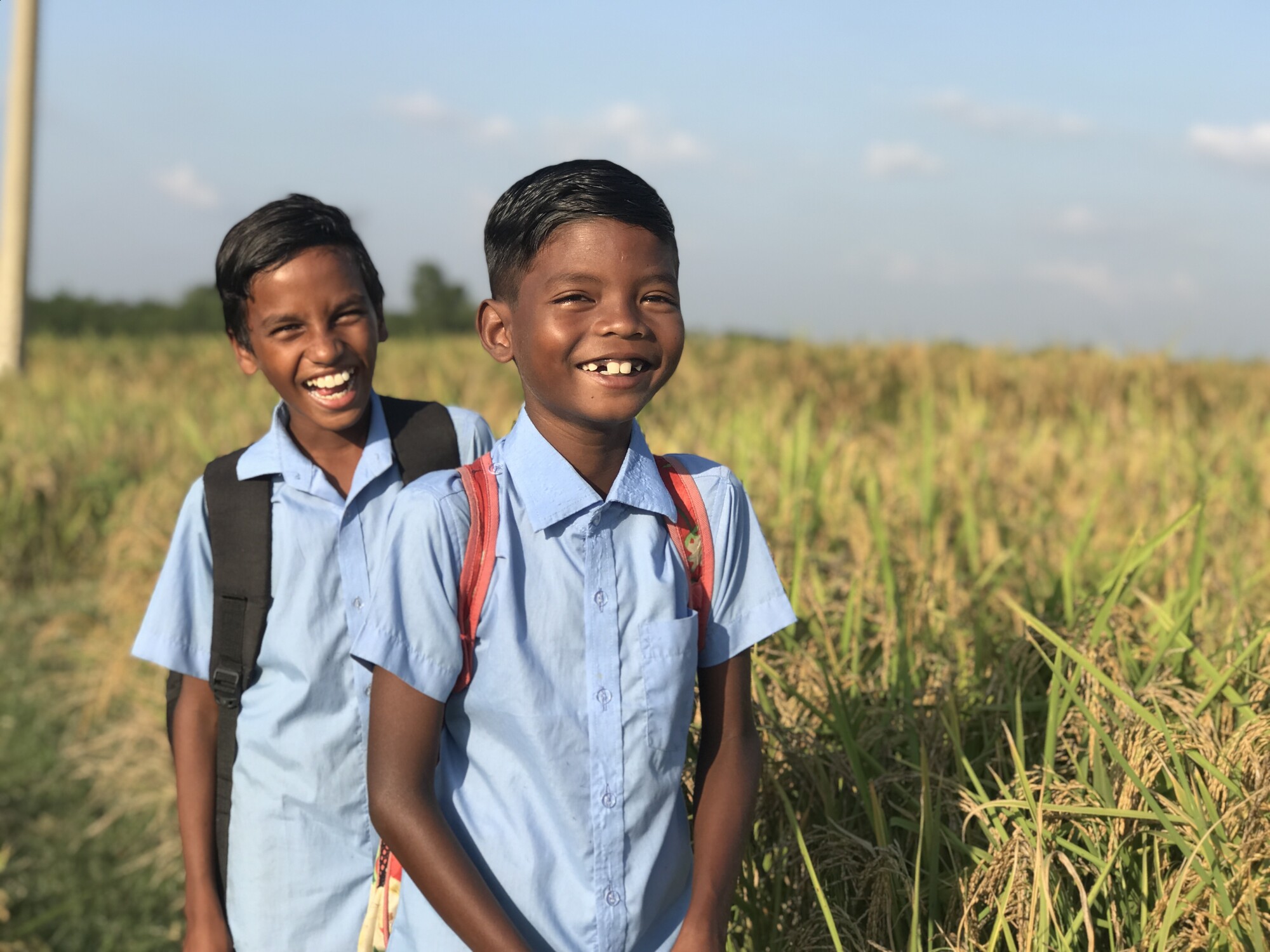 two smilingboys wearing blue button-down shirts and backpacks stand in a grassyfield