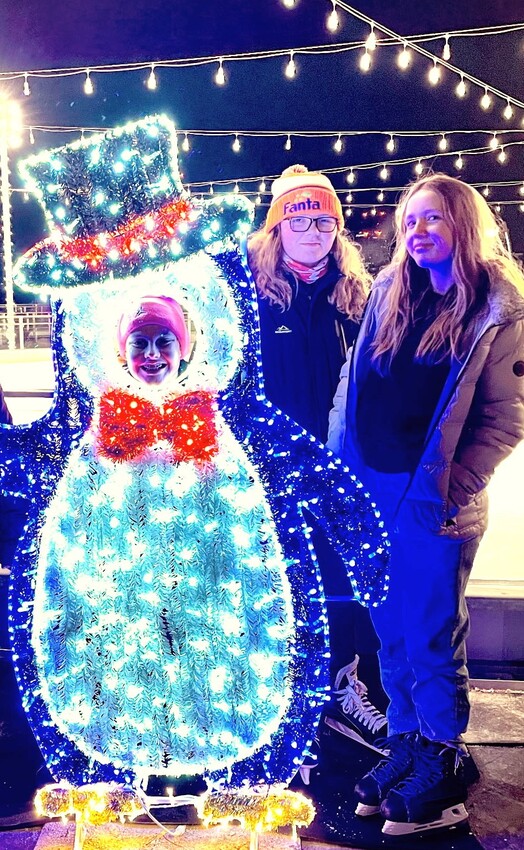Three individuals stand near a lit penguin decoration. One person is inside the penguin, while the others pose beside it.