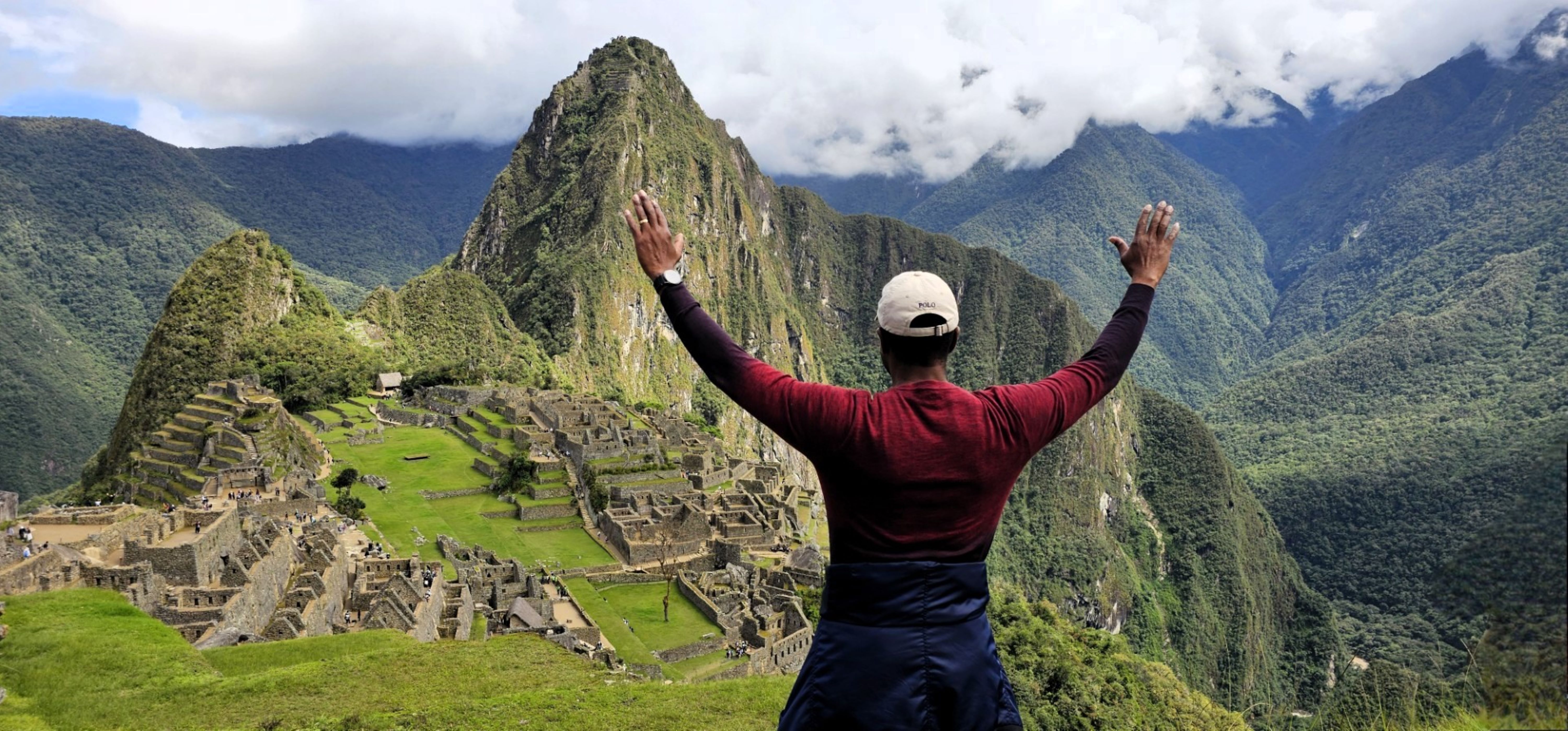 a man stands facing away from the camera with hands raised to the sky in front of Machu Picchu